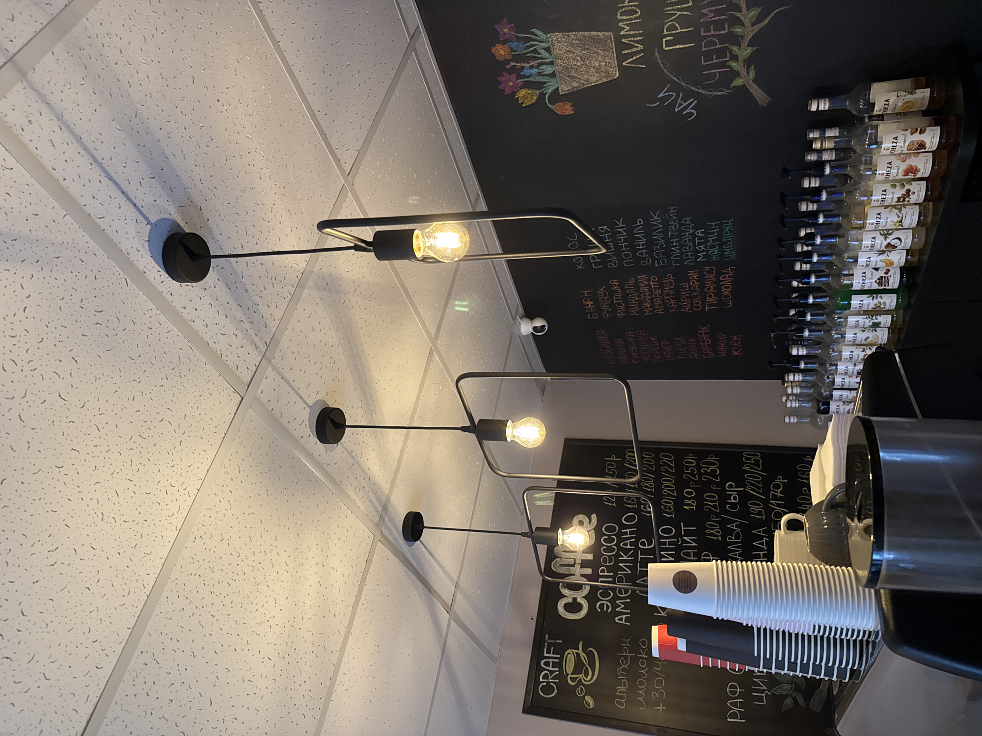 Here is a description of the image:
The image shows a cafe or coffee shop setting. The scene is shot from a low angle looking up towards the ceiling and wall. The ceiling is made of white acoustic tiles. Modern, black wall-mounted light fixtures with exposed Edison bulbs are attached to the ceiling.
The wall is painted black and features a menu written in chalk. The menu includes descriptions of various drinks (espresso, americano, latte) and flavorings (strawberry, banana, hazelnut, raspberry, etc.). There are also bottles of syrups lined up on a shelf above the menu. A stack of disposable coffee cups is visible on a counter next to a coffee grinder, which is out of focus due to the close proximity.