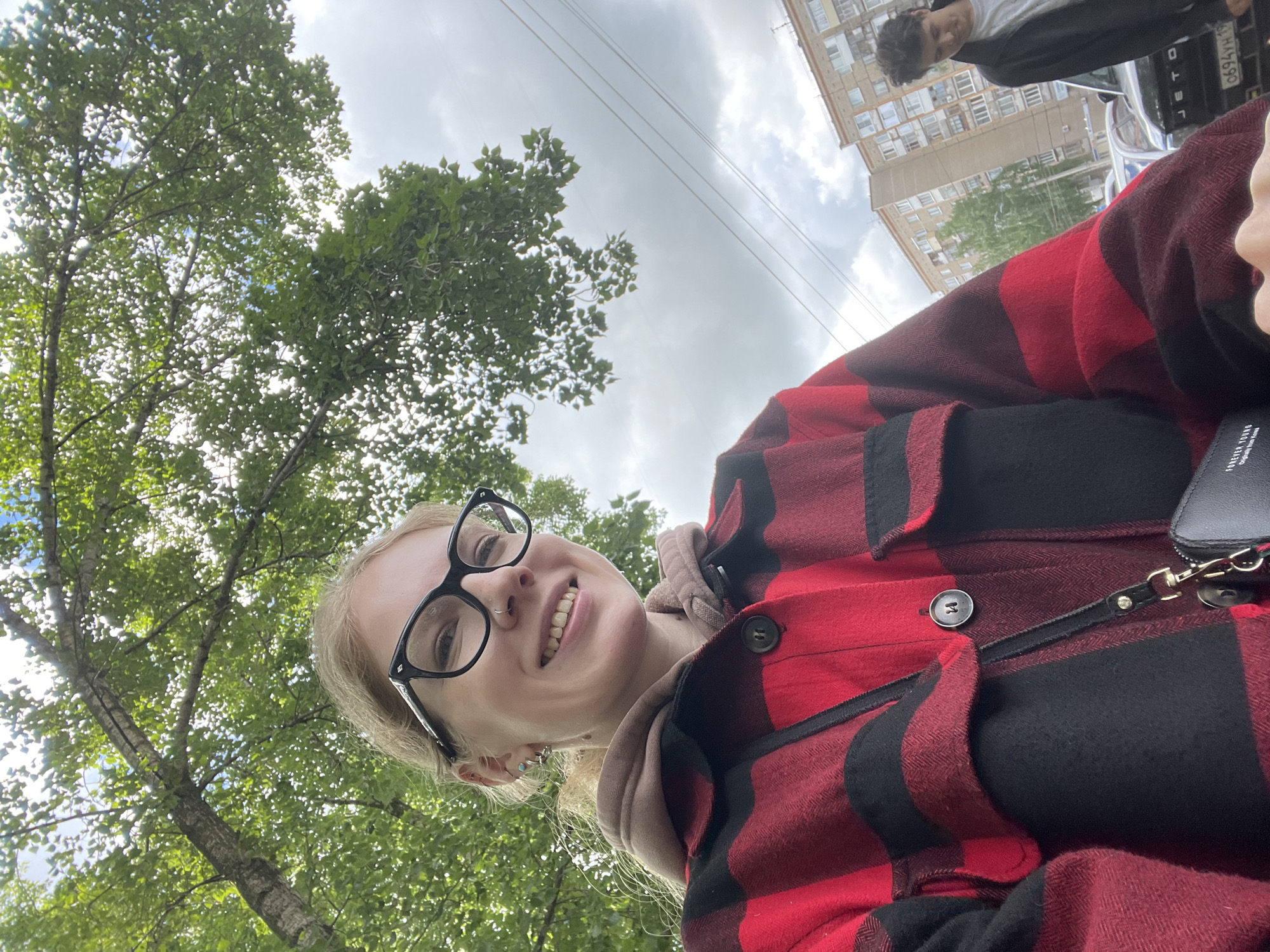 A low-angle photograph shows a smiling woman wearing glasses and a red and black checkered jacket. Trees with green leaves fill the upper left corner of the frame, while a building is visible in the upper right. Part of a car and a person are also in the upper right. Power lines run across the sky. Based on license plates visible in the image, the location is Izhevsk, Russia.