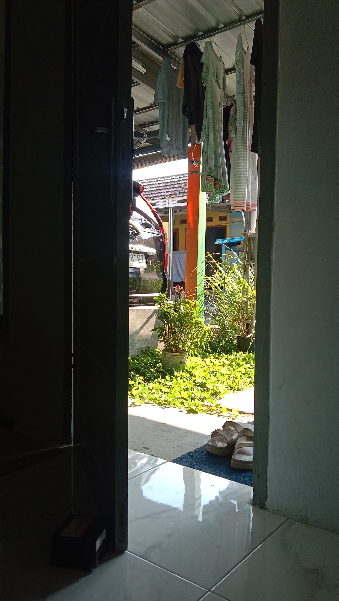 The image depicts a view through an open doorway into an outdoor area in Karangmangu, Indonesia. In the foreground, on the left, a dark, shadowed doorway frame is visible, with a dark object that appears to be a small box or container resting on the floor near the bottom left corner. The floor is tiled with large, light-colored tiles that reflect the bright sunlight.
In the middle ground, beyond the doorway, a clothesline is strung under a corrugated metal roof, with several items of clothing hanging on it. A dark-colored car is parked behind a concrete wall or raised platform. Green plants are growing in pots and on the ground in front of the car and wall. The ground is covered with a low-lying green plant that appears to be some type of ground cover.
To the right of the image, a blue doormat is placed on the tiled floor, with a pair of light-colored sandals resting on it. The environment appears to be a residential area, likely a backyard or front yard. The lighting suggests it is daytime, with bright sunlight casting strong shadows. The overall impression is a casual, everyday scene.