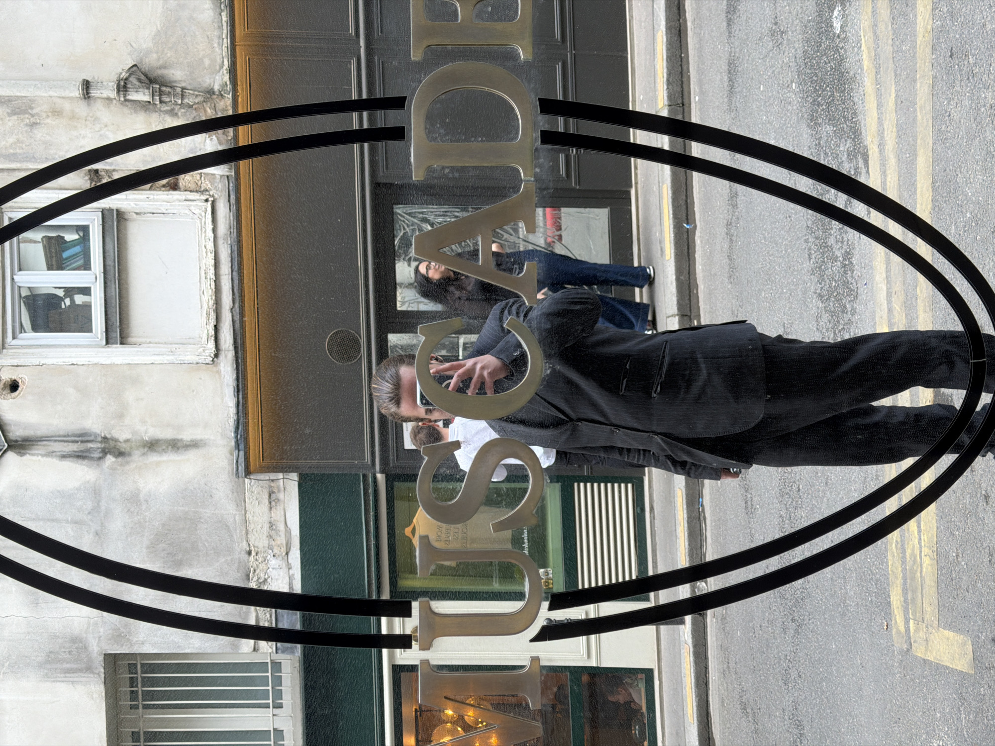 The image depicts a reflective shop window at the "MUSCADE" boutique, located at 26 Rue des Francs Bourgeois, 75003 Paris, France. Gold-colored, metallic letters spelling "MUSCADE" are affixed to the glass, along with two curved, vertical black metal strips that create an oval framing effect.
Centered in the reflection is a male individual, appearing to take a selfie with a dark-colored smartphone held up to his face, partially obscuring it. He has dark hair pulled back and wears a dark pinstriped suit. To his immediate left, the reflection of a second male in a white shirt is partially visible. Further back and to the right, a woman with dark hair and glasses, dressed in a dark top and blue jeans, stands on the sidewalk.
The reflected background reveals a narrow Parisian street lined with multi-story, aged stone and plaster buildings. These structures feature various windows, some with security bars, and exhibit visible signs of wear, including cracks and discoloration. The asphalt street below has faded yellow markings. Through the gaps in the "MUSCADE" lettering and around the reflections, parts of the shop's interior are discernible, showing warm lighting and a light-colored t-shirt on display, visible in reverse. The scene is illuminated by ambient daylight, suggesting an overcast day.