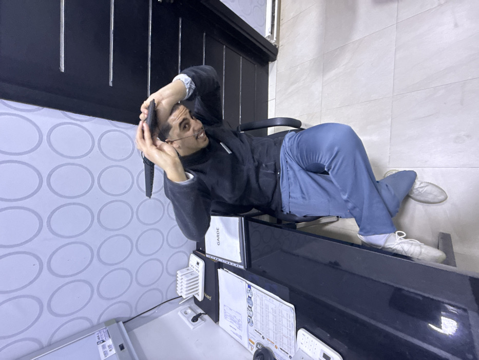 A male individual is seated in a black office chair behind a dark, glossy desk in an indoor environment in Meknes, Morocco. The man wears a dark long-sleeved top over a light-cuffed undershirt, light blue trousers, and mismatched light-colored shoes. His left foot wears a laced athletic shoe, while his right foot wears a slip-on shoe. His hair is dark and short, and he has a mustache and goatee. He is looking directly forward with an open-mouthed expression, displaying teeth. His hands are raised above his head, holding a black rectangular object, possibly a smartphone, horizontally. A thin dark wire or object is visible near his right cheek.
The desk surface contains various items: a white remote control, multiple sheets of paper with text, a white multi-slot device resembling a pager charging station, and a black folder labeled "GARDE." A white electrical outlet with a black cord plugged in is visible on the light-colored wall directly above the desk. This wall is covered in wallpaper featuring a repeating pattern of light grey concentric circles. To the right of the seated individual is a dark, vertically grooved panel, possibly a door or wall feature. The floor consists of light-colored, large square tiles. The setting suggests an office or reception area.
