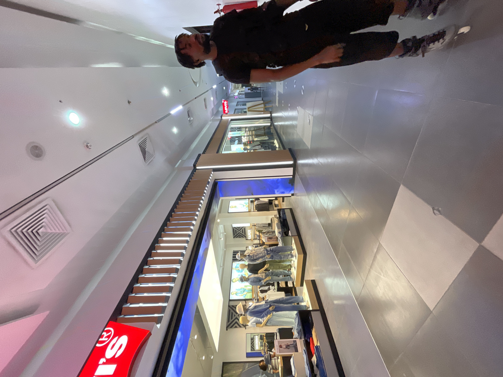 A man wearing a black t-shirt, dark shorts, and rollerblades stands in a brightly lit indoor mall hallway, looking to his right. The hallway features a modern design with a checkered tile floor, illuminated by overhead lights and light emanating from storefronts. To his left, a Levi's store is visible, displaying mannequins dressed in denim attire. The store's red and white logo is prominent. The environment appears clean and well-maintained, typical of a commercial shopping area in Moba, Nigeria.