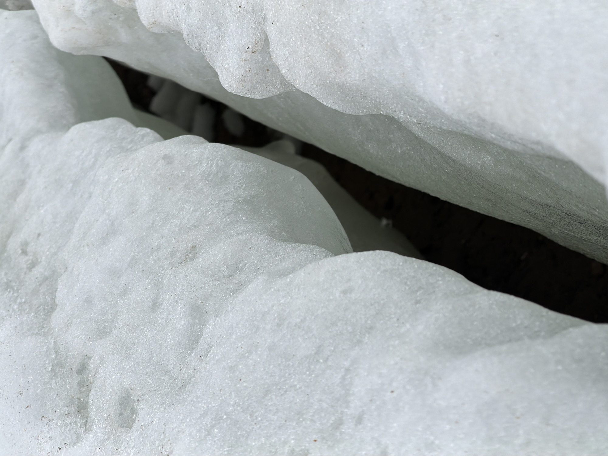 The image shows a close-up view of ice or snow formations, potentially near or over a body of water, creating a dark crevice between the snow masses. The scene lacks specific details identifying Chaikovsky, Russia, or any people, objects, or interactions. The focus is solely on the textured surface of the snow and the dark void it creates.