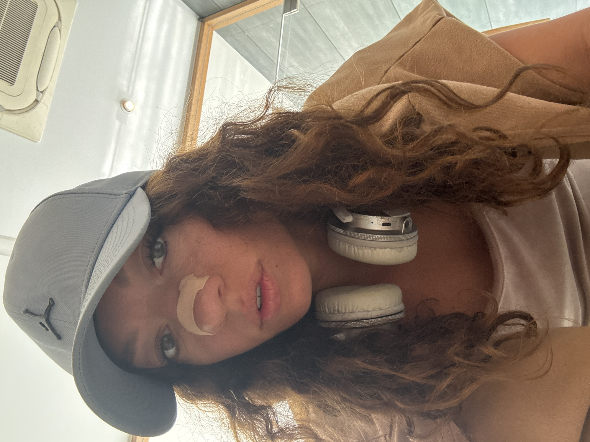 A selfie shows a woman with long, curly, brown hair wearing a gray baseball cap with a black Jordan logo and beige headphones around her neck. There is a bandage on her nose. The location is likely indoors, as the background features a ceiling with an air conditioning unit and a window frame. The image provides no evidence that the photo was taken in Đà Nẵng, Vietnam.