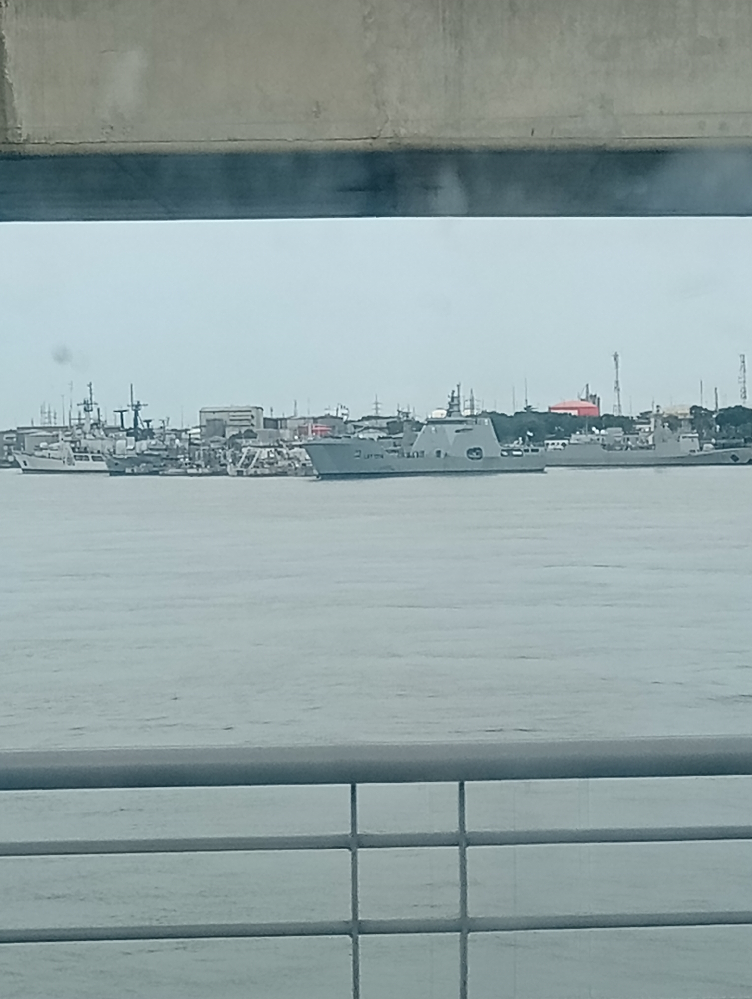 The image shows a waterfront scene in Lagos, Nigeria, likely viewed from an elevated position such as a bridge. In the foreground, a light-colored railing is visible. Beyond the railing is water, which appears calm. Further in the distance, several naval vessels are docked. The skyline includes buildings, cranes, and other industrial structures. The overall scene is overcast.