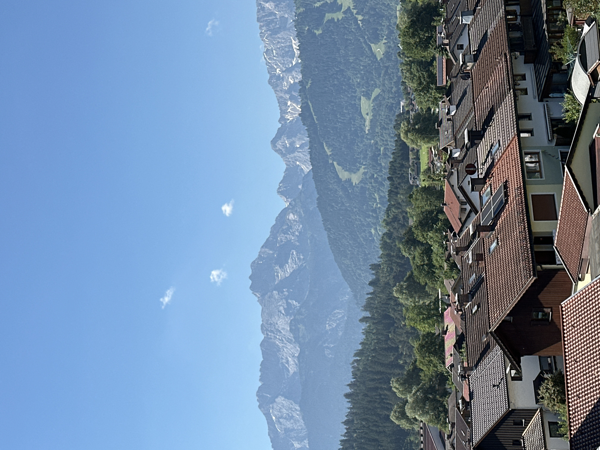 This is a vertical, medium-shot photograph of a town with mountains in the background. The sky is clear and blue with a few small clouds. In the foreground, there is a dense collection of houses with red tile roofs. Some houses have solar panels on their roofs, and chimneys are visible on many of them. The houses are packed closely together, forming a residential area.
Behind the houses, a lush green forest stretches across the middle ground. The trees are dense and vary in shade, indicating a mix of evergreen and deciduous species. The forest appears to cover rolling hills, and a ski slope with cut-out paths is visible on one of the hillsides, suggesting the area is used for winter sports.
In the background, majestic, jagged mountains rise up, their peaks partially covered with snow. The mountains have a rugged, rocky appearance, and their lower slopes are also covered with forests. The overall impression is a scenic view of a town nestled at the base of a mountain range. The scene depicts the town of Garmisch-Partenkirchen, Germany, a popular tourist destination known for its alpine scenery and winter sports. No people are visible in the image.