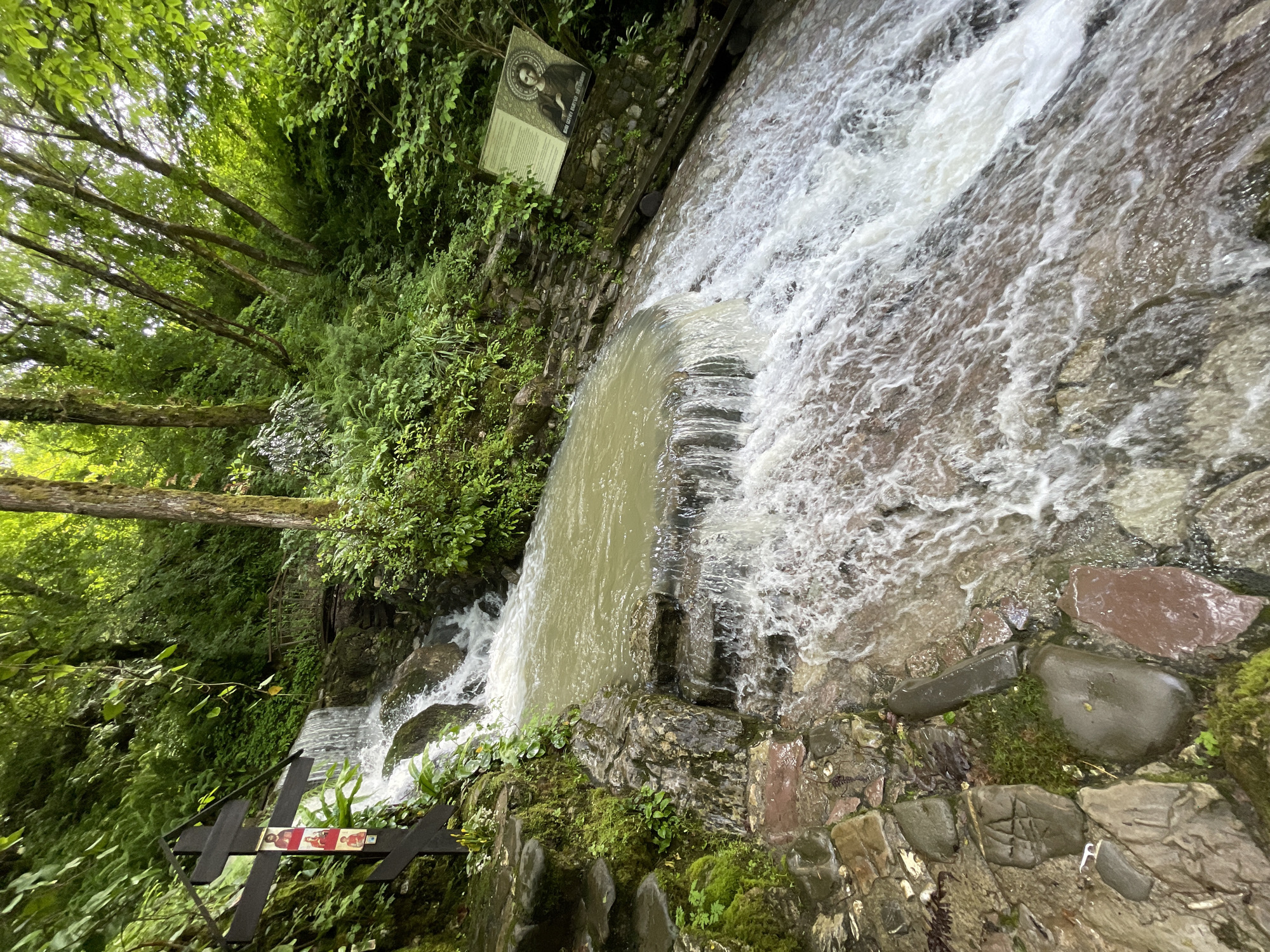 The image depicts a waterfall located in Solokh-Aul, Russia. The waterfall flows over a series of stone steps and rock formations covered in moss. Lush green vegetation surrounds the waterfall, including trees and various plants. A wooden cross with religious images is positioned near the base of the waterfall on the left side. Higher up on the left side, a sign with a portrait and text is affixed to the stone wall. The water appears turbulent and fast-moving. The scene suggests a natural setting within a developed area, indicated by the constructed stone structures.