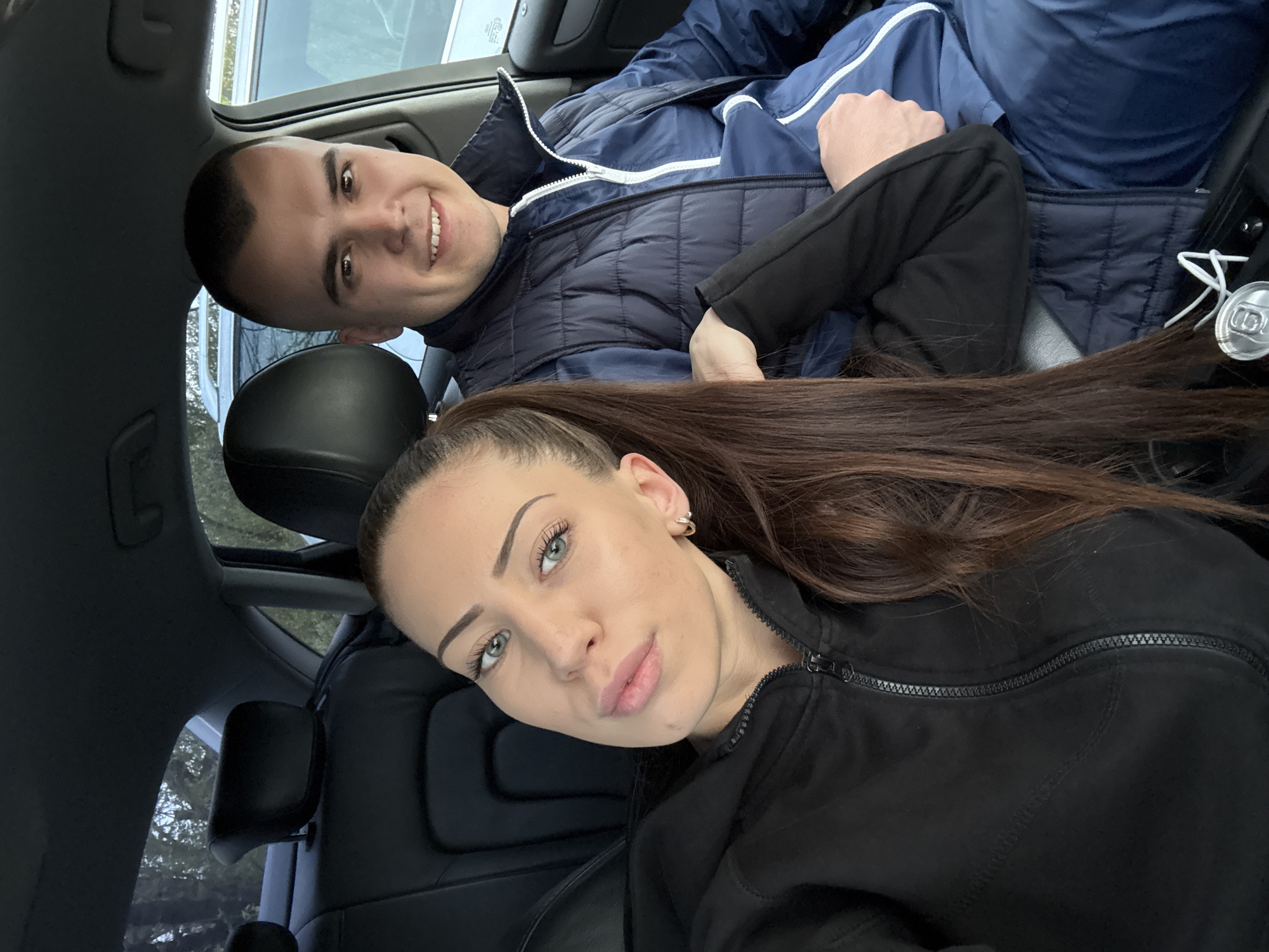 The image shows a man and a woman inside a car. The man is wearing a navy blue puffer jacket with white accents and is smiling. The woman is wearing a black jacket and has long, dark brown hair. There is a soda can visible near the man. The interior of the car appears to be black leather. A sunroof is visible above. The location is unknown.