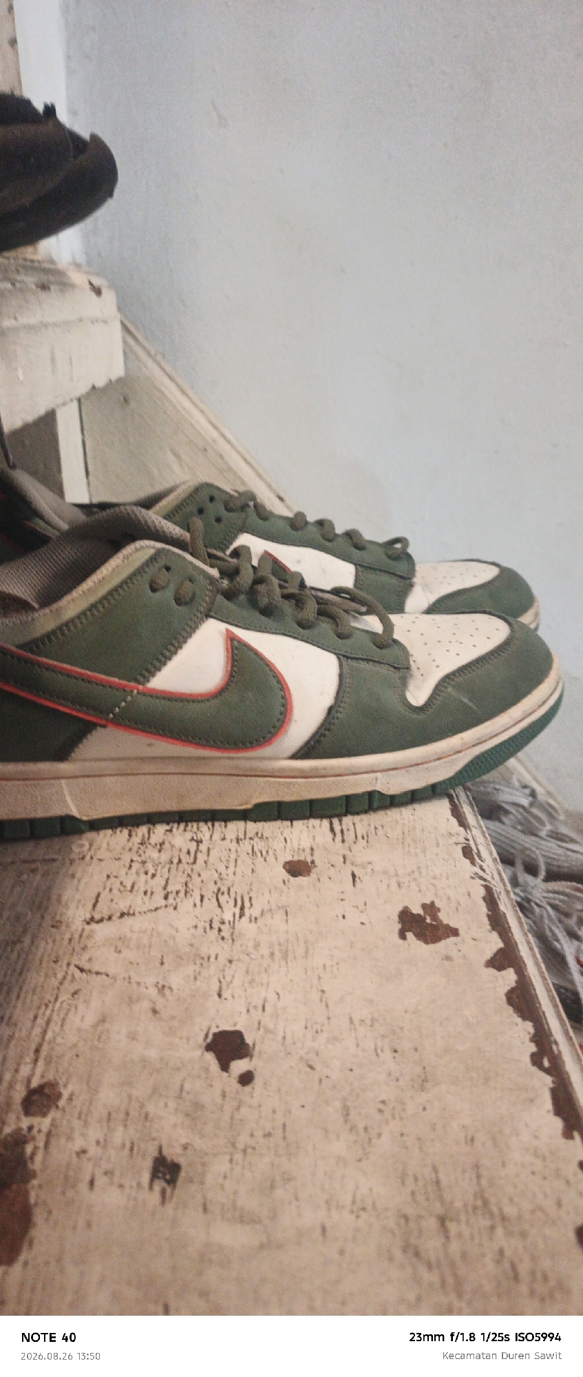 The image displays a pair of dark green and white Nike Dunk Low sneakers with red accents, positioned on a worn, distressed wooden surface. The sneakers appear to be used, with visible signs of wear and tear on the soles and uppers. The background is out of focus, suggesting a shallow depth of field, and shows a textured wall and a portion of a dark shoe at the top left. The bottom of the image includes text overlays indicating "NOTE 40," a date and time stamp "2026.08.26 13:50," and camera settings "23mm f/1.8 1/25s ISO5994" along with a location "Kecamatan Duren Sawit." Kecamatan Duren Sawit is an administrative district located in East Jakarta, Indonesia.
