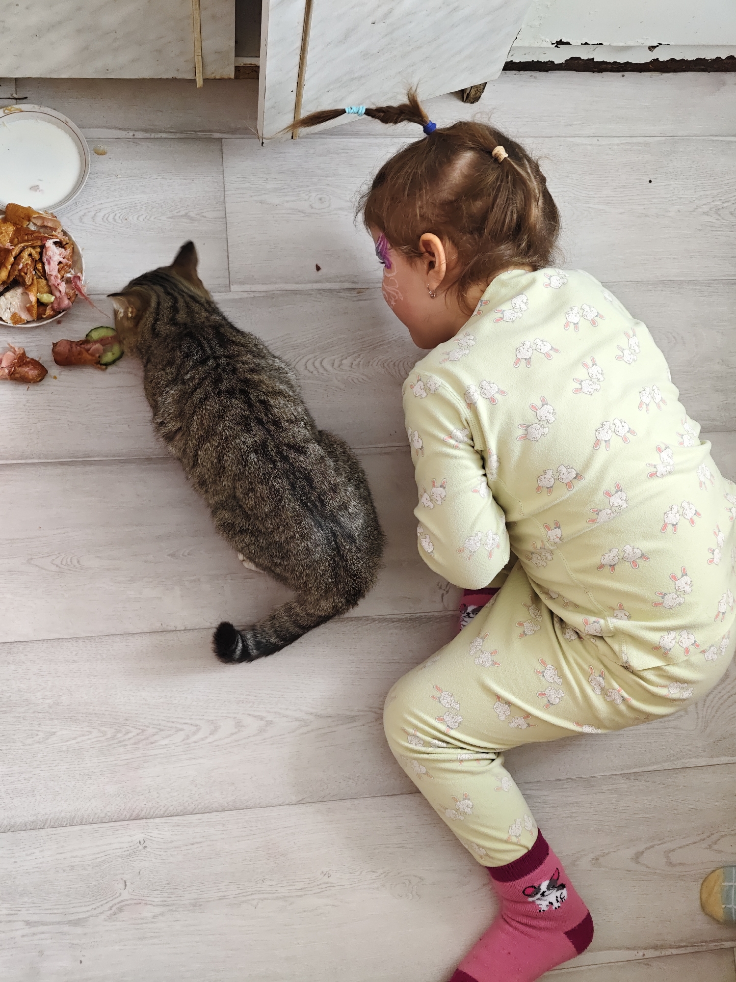 A young child wearing yellow pajamas with a bunny print and pink socks is kneeling on a light-colored wood floor, looking at a tabby cat. The cat is standing next to a plate of food scraps, including what appears to be chicken bones, sausage, and cucumber slices. The background includes a white cabinet with peeling paint. The location is potentially in the city of Уральское, Russia.