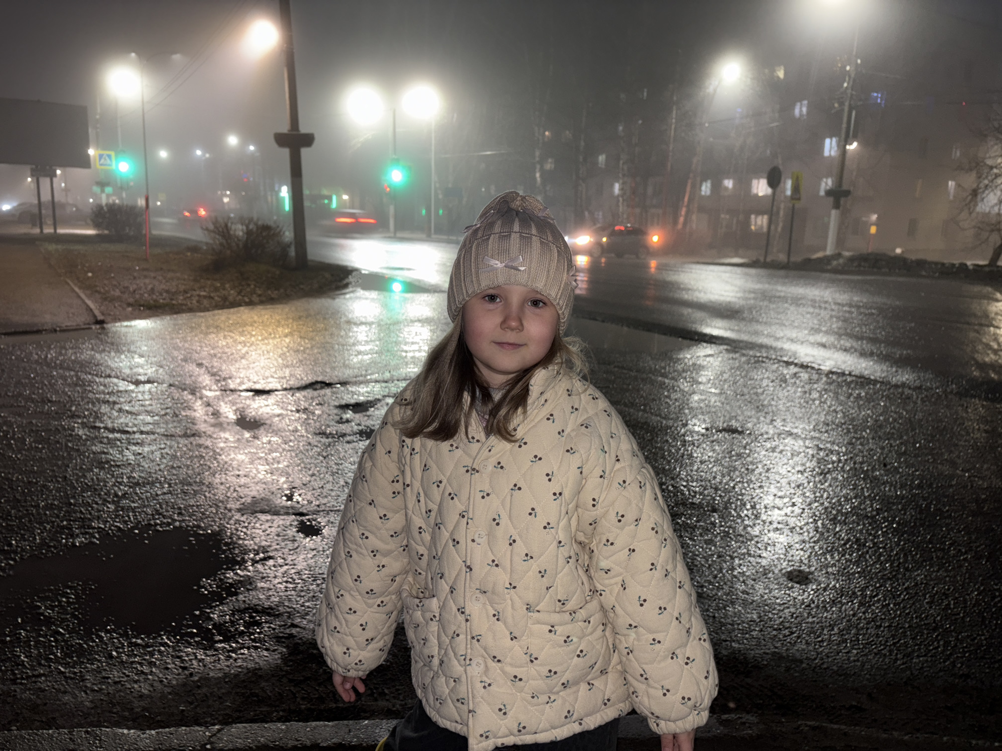 A young girl stands on a wet pavement in the city of Chaikovsky, Russia. She is wearing a light-colored quilted jacket with a small cherry pattern and a knitted hat with ribbon accents. The street is wet, reflecting light from the streetlights. Streetlights and traffic signals are visible in the background, and cars are blurred in motion. Fog obscures the buildings and trees in the distance.