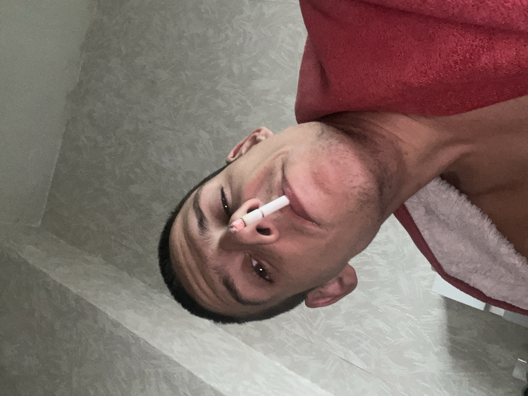 A man with short dark hair and brown eyes is holding a lit cigarette in his mouth. He is wearing a red and white hooded bathrobe. The background appears to be a textured grey wall. The image is taken at an angle. There is no indication of the location being Sofia, Bulgaria.