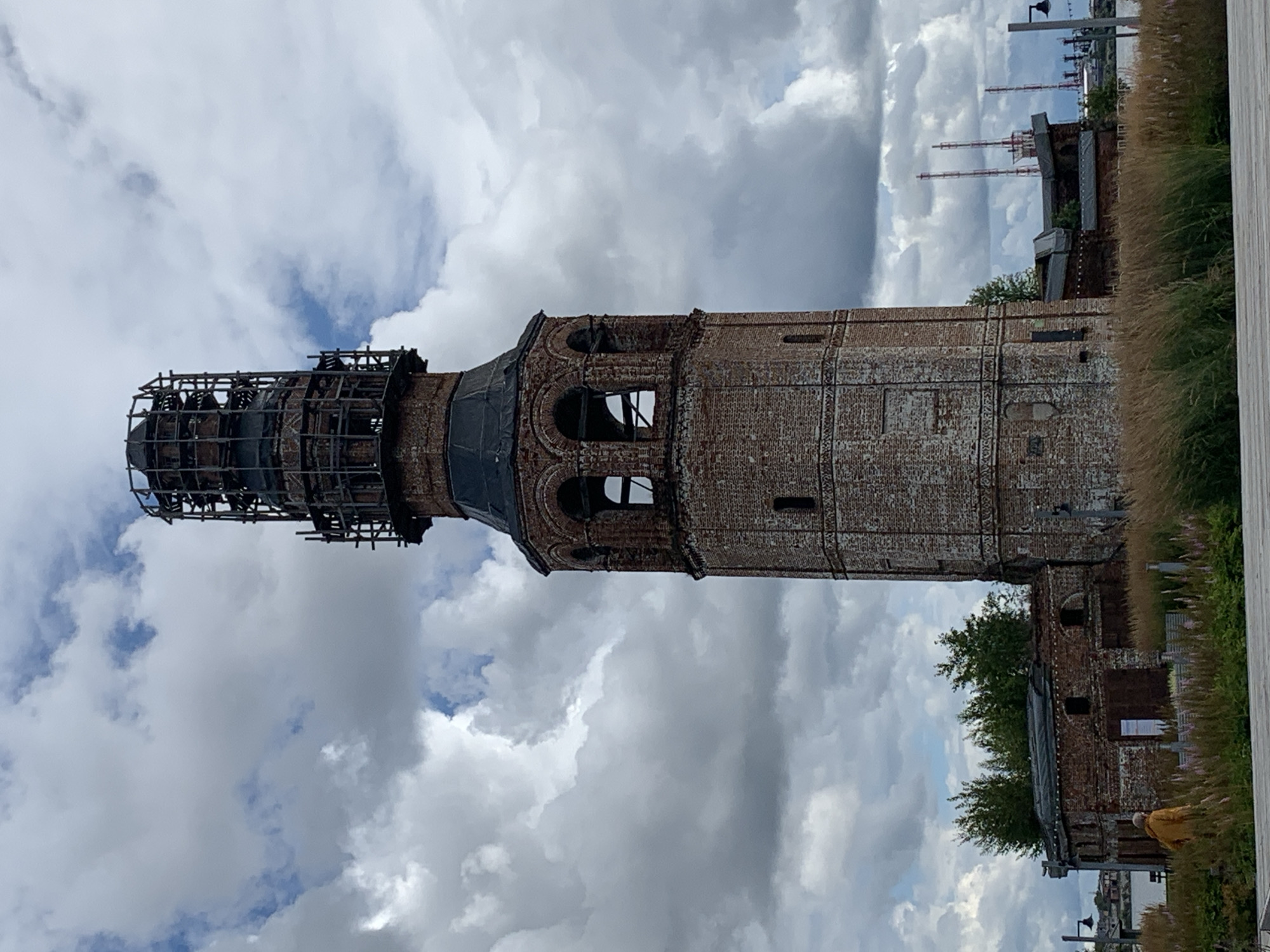 The image depicts a tall, octagonal brick tower, appearing weathered and possibly undergoing renovation or in a state of disrepair. The top of the tower is encased in a metal scaffolding structure. Below the scaffolding, the tower features arched openings, reminiscent of a belfry, and various brickwork details. Attached to the base of the tower is a lower, brick building, also showing signs of age. In the foreground, tall grasses and some flowering plants are visible. A person wearing a yellow jacket is walking through the grass. In the background, several industrial structures with tall stacks and cranes are visible, suggesting an industrial area or port. The sky is filled with dramatic, cloudy formations. The scene is identified as being in the city of Usolye, Russia.
