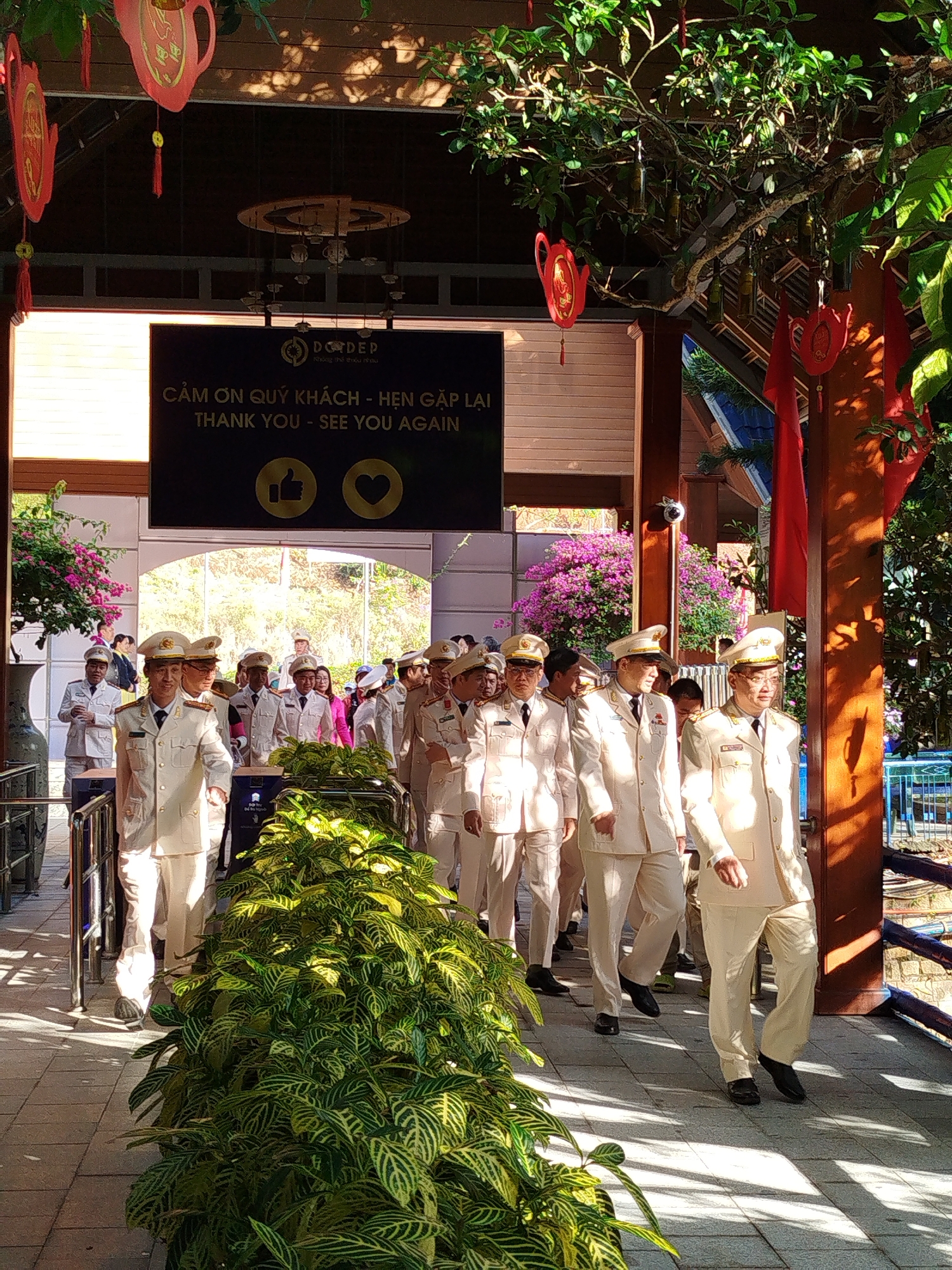 A group of people dressed in similar white uniforms with caps walk through a partially shaded passage, seemingly outdoors, in Da Lat, Vietnam. A sign overhead, in both Vietnamese and English, reads "CẢM ƠN QUÝ KHÁCH - HẸN GẶP LẠI THANK YOU - SEE YOU AGAIN." The sign includes a thumbs-up and heart icon. The pathway has a light-colored stone surface. A large green potted plant is to the left of the group. Overhead, decorations including red teapot-shaped ornaments and red flags can be seen. The scene appears to be a welcoming or farewell gesture at a public place in Da Lat.