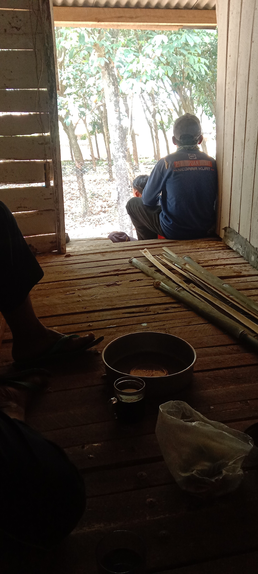 The image depicts a rustic wooden porch or dwelling with a view of trees and foliage outside. In the foreground, a person's leg and foot, wearing a sandal, are visible on the wooden floor. To the right, a large metal bowl and a small glass containing a dark liquid are placed on the floor. Several bamboo poles are also stacked on the wooden planks.
Further back, a person wearing a dark blue long-sleeved shirt with lettering on the back and a dark cap is seated, facing away from the camera and looking outwards. Another individual, possibly a child, is seated in front of the person in the blue shirt. The background shows a fence and a natural outdoor environment with trees.
The scene suggests a relaxed, possibly informal setting. The presence of bamboo poles and the rustic construction of the dwelling might indicate a rural or semi-rural location. The inscription on the shirt, "PEREMPUAN HEBAT NEGARA KUAT," translates to "Great Woman, Strong Nation" in Indonesian, suggesting a patriotic or nationalist theme.
While the image itself does not provide direct information about the city of Utama Jaya Mataram, Indonesia, the inscription on the shirt indicates a connection to Indonesia. If this location were indeed Utama Jaya Mataram, it would imply a setting within that area. The overall atmosphere is subdued, with natural light filtering in from the open doorway.