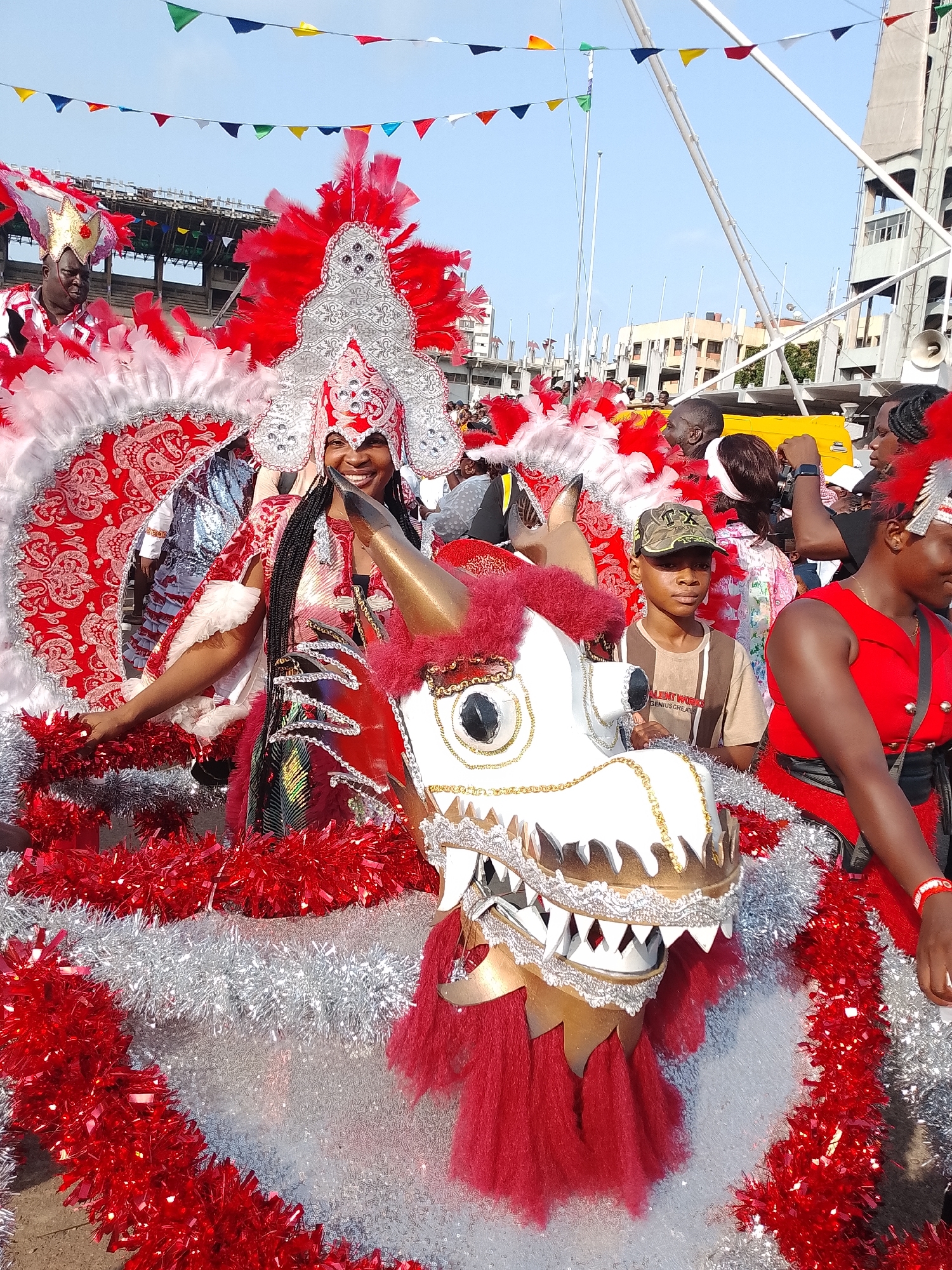 A medium, outdoor shot shows a large group of people in carnival dress in Lagos, Nigeria. The main subject is a woman in the center, wearing a tall red and silver headpiece and a matching red and silver dress with white feather trim. She is smiling. In front of her is a white dragon head with gold trim, also decorated with red tinsel. A young boy in a camouflage cap is standing beside the dragon head. A woman in a red dress is on the right side of the frame. Behind the main subject, there are other people in carnival attire, as well as other spectators. Colorful pennants are strung overhead. A stadium-like structure is visible in the background. A light blue sky is above.