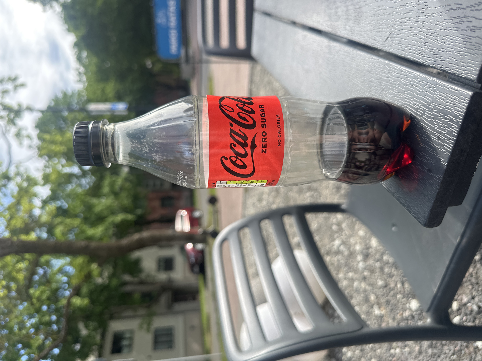 A close-up, eye-level shot shows a transparent plastic bottle of Coca-Cola Zero Sugar with a black cap. The bottle is partially filled with dark brown liquid and rests on a textured, dark gray outdoor table. The Coca-Cola Zero Sugar label, predominantly red with white lettering, is clearly visible. Behind the bottle, a gray outdoor chair with slatted seating is positioned to the left. The background is softly blurred, revealing an outdoor setting with green trees and a white building. A red car is visible in the distance, and a blue sign with white text is present further back. The ground appears to be covered in gravel. The scene is likely an outdoor seating area of a cafe or restaurant in Tallinn, Estonia, given the architectural style of the building and the presence of typical outdoor furniture.
