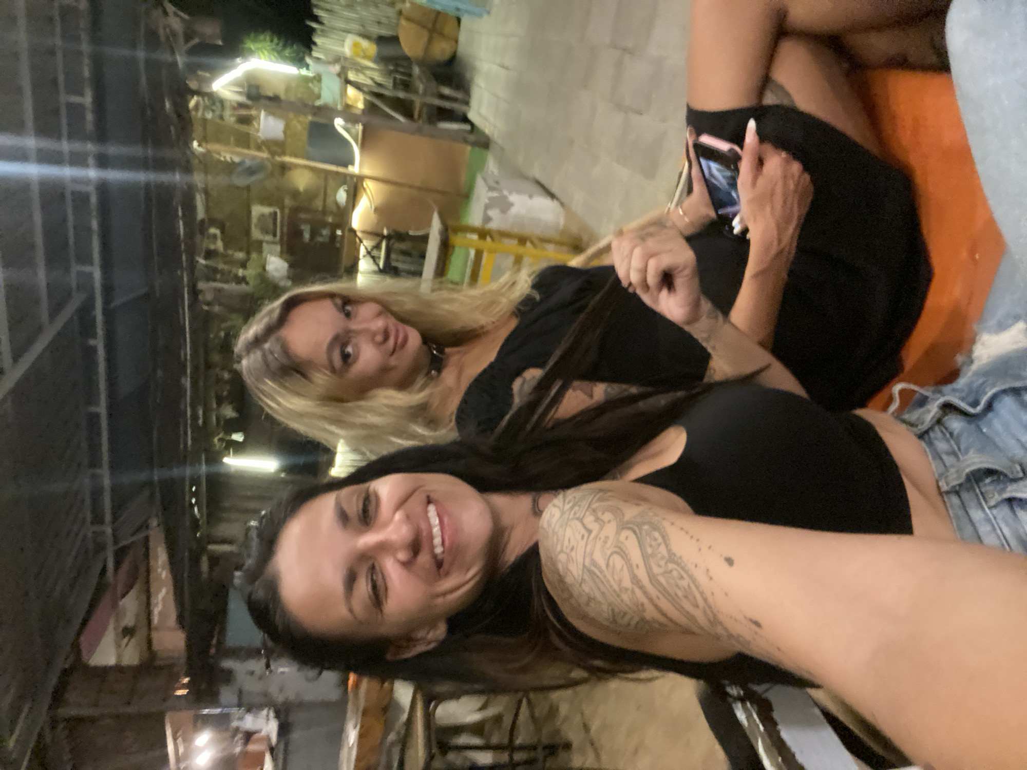 The image captures two women seated outdoors at what appears to be a casual establishment in Mũi Né, Vietnam. The woman in the foreground, on the left, has dark hair and is smiling broadly. She has extensive tattoos on her left arm and shoulder. She is wearing a black tank top and denim shorts. The woman in the background, on the right, has blonde hair and is wearing a black dress and a choker necklace. She is holding a smartphone in her right hand. The setting suggests an outdoor seating area with a rustic, possibly beach-themed ambiance, indicated by the wooden structures and lighting in the background.