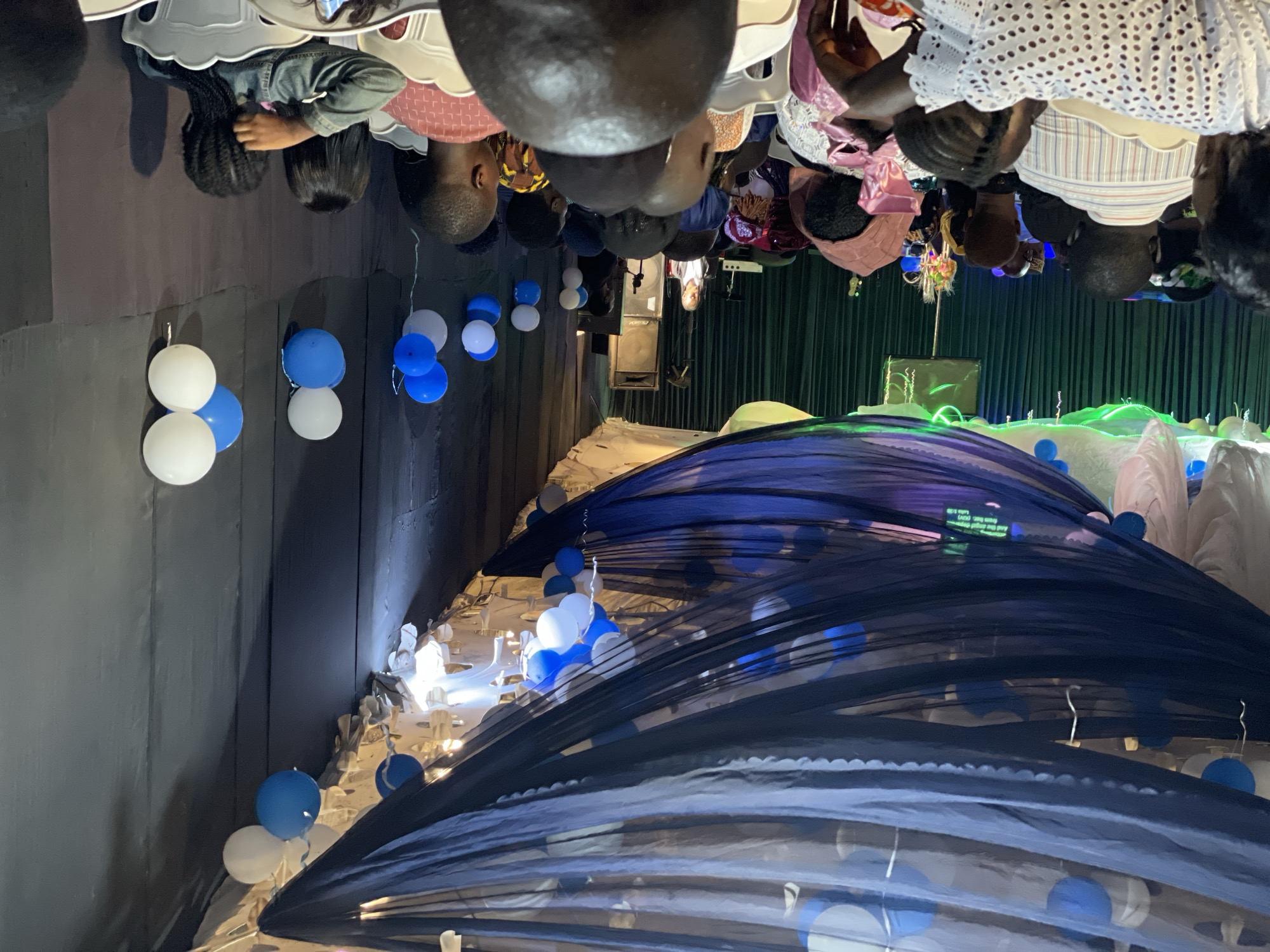 The image depicts an indoor gathering, possibly a church service or event, in the city of Wumat, Nigeria. The space is decorated with blue and white draped fabric suspended from the ceiling, along with clusters of blue and white balloons. A stage is visible at the far end of the room, with a woman standing at a podium. The audience is seated in rows of white chairs, facing the stage. The attendees appear to be of various ages and are dressed in casual and formal attire. The lighting is dim, with spotlights illuminating the stage area. The overall atmosphere suggests a celebratory or formal occasion.