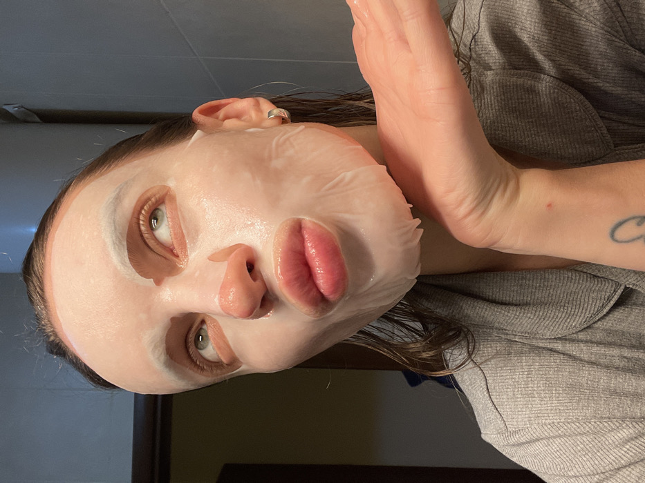 A fair-skinned person wearing a sheet mask poses sideways. The mask covers most of the face, with openings for the eyes, nose, and lips. The eyes are light-colored. A hand is raised to the side of the face. The person is wearing a gray, textured shirt. A partial tattoo is visible on the arm. The background is dark, with hints of a wall.