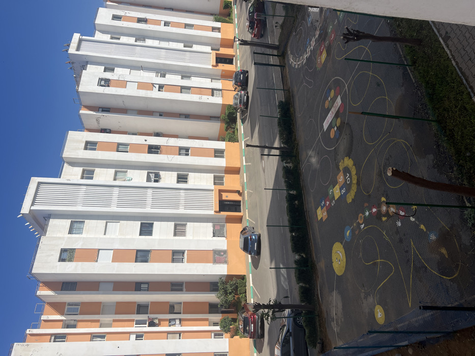 A high-angle view captures a tall, white building with orange accents under a clear blue sky. Parallel to the building runs a road with parked cars. Adjacent to the road is a playground with colorful markings including a hopscotch and various circular designs, bordered by greenery. The composition is oriented vertically, showcasing the building's height and the playground's layout.