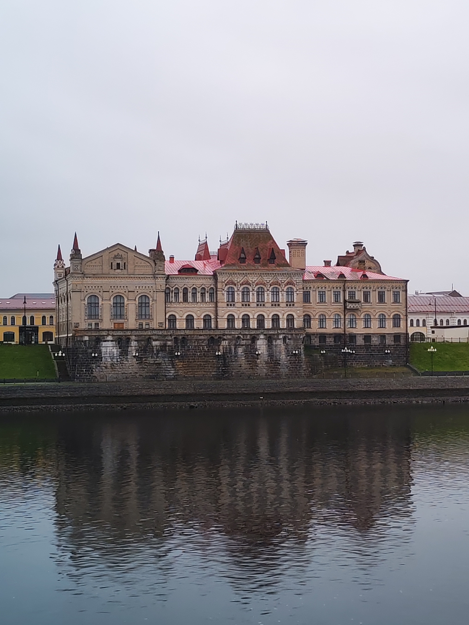 The image depicts the Palace of Agriculture in Bol'shie Mkhi, Russia, reflected in a body of water. The building has a light-colored facade with red roofing and decorative spires. It stands on a stone base along the waterfront. Adjacent buildings and landscaping are visible to the sides. The sky is overcast. The water reflects the building and sky.