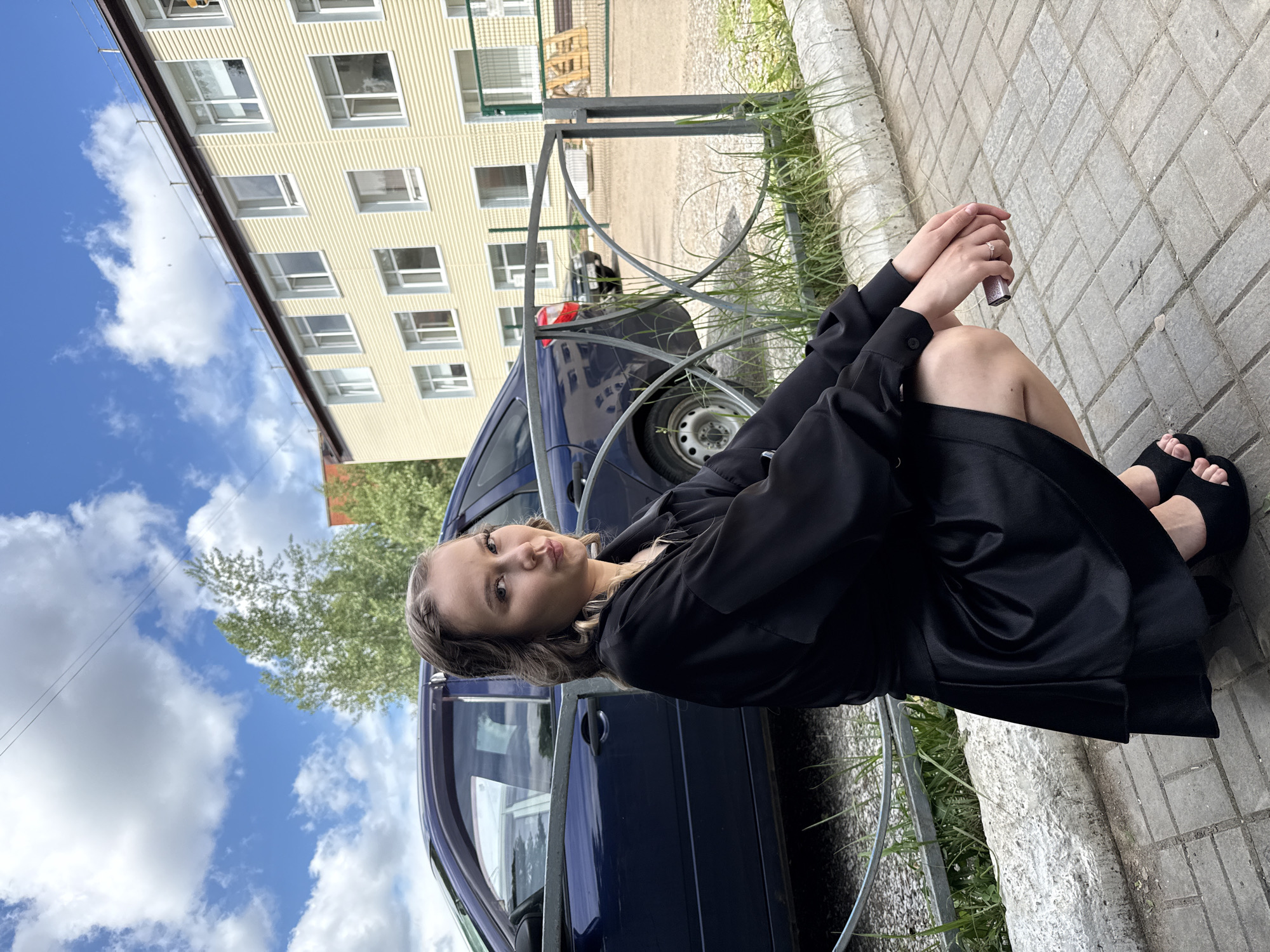 A young woman is sitting on the sidewalk next to a blue car. She is wearing a black dress and black sandals. She has blonde hair and is looking at the camera with a slightly pouted expression. Her hands are clasped in her lap, and she is wearing a ring on her finger. The car is parked on the street, and there is a yellow building in the background. The sky is blue with white clouds. The weather appears to be sunny and warm, likely daytime. The scene appears to be in an urban or suburban environment, possibly in Chaikovsky, Russia, given the provided location context. There is no visible text in the image.
