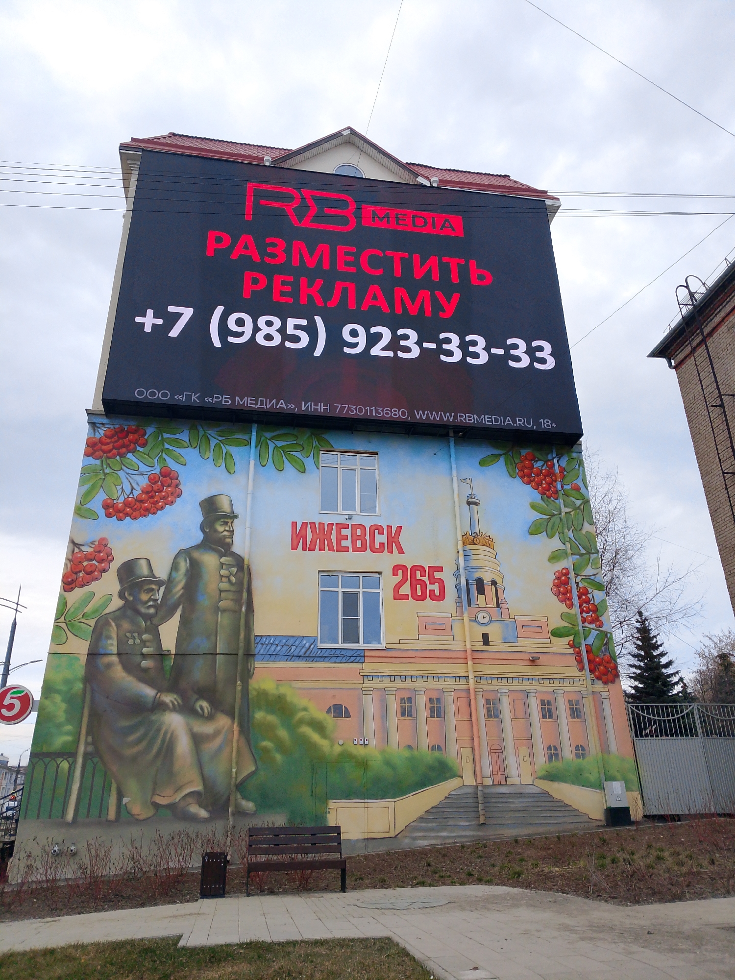 The image shows a building in Izhevsk, Russia, with a large advertisement on top and a mural on the side. The advertisement, in red and white letters, says "RB MEDIA," "Place an Ad," and the phone number "+7 (985) 923-33-33." Below the advertisement is the mural, featuring a depiction of two men in period attire, likely historical figures, and a representation of the Izhevsk city hall or a similar landmark. The word "Izhevsk" is written in red. Also, in red, is the number 265. The mural also depicts rowan berries on green stems surrounding the building. In front of the building, there is a bench and some sparse vegetation. A sign with the number 5 in a red circle can be seen to the left, along with a street light. A brick building is visible to the right of the painted building. The sky is overcast.
