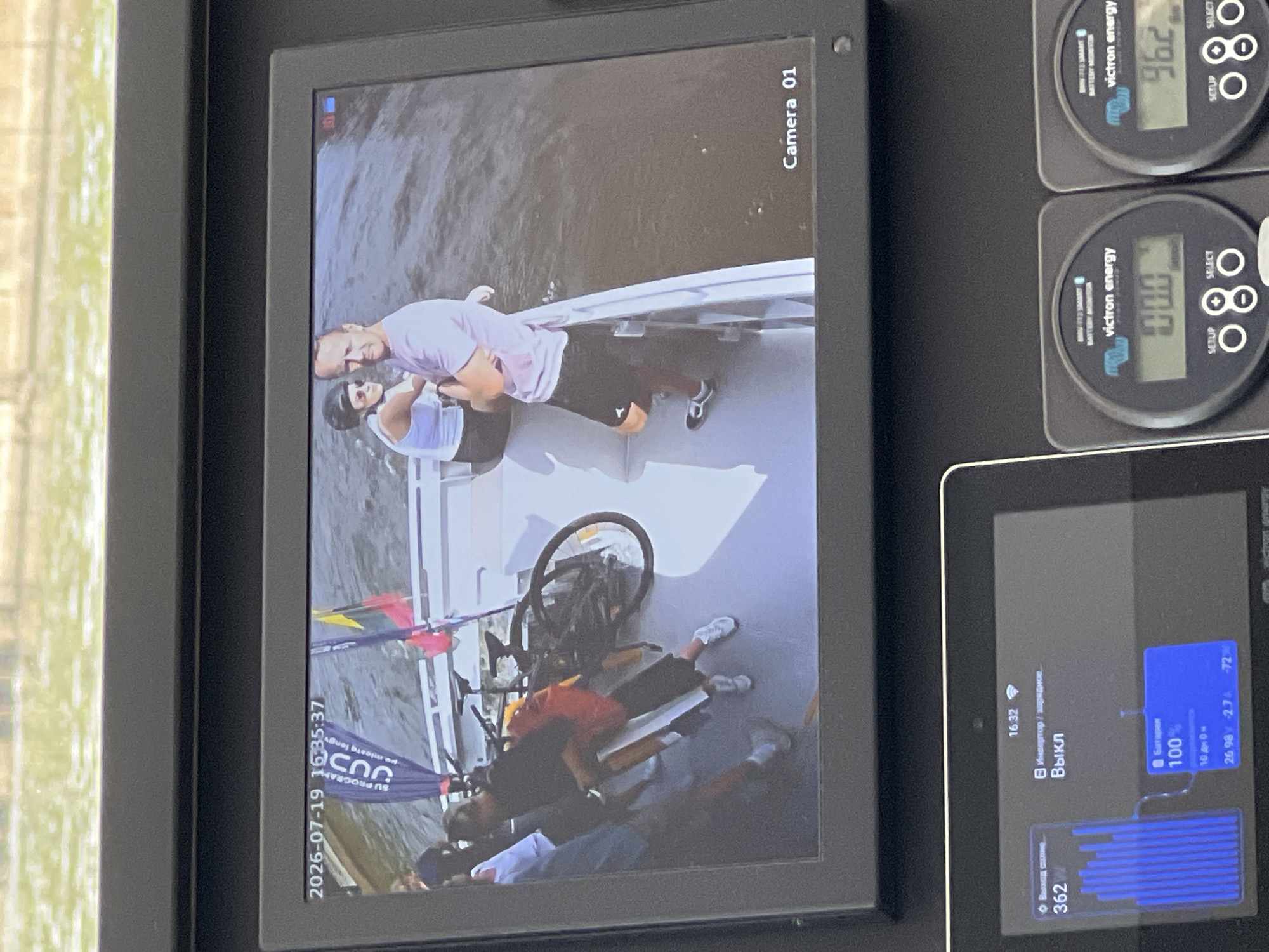 On July 19, 2026, at 16:35:37, a security camera monitors an event on a boat. Two individuals, a man and a woman wearing a helmet, are visible on the deck. The man is wearing a pink t-shirt and dark shorts, while the woman is wearing a white tank top and dark shorts. A bicycle is also present on the boat, leaning against a railing. In the background, two flags are visible, one blue and one yellow and red. The boat appears to be on a body of water, with the cityscape of Vilnius, Lithuania, potentially visible in the distant background. The camera displays "Camera 01" in the lower right corner. Below the main monitor, a digital tablet displays system information. It shows the time as 16:32, with indicators for "Output current: 362W" and "Inverter/charger: Off". A battery status indicator shows "Battery 100%" with "10 days 0h" remaining and readings of "26.98 -2.7 -72". Two Victron energy battery monitors are also visible, one displaying "0.00" and the other "96.2".