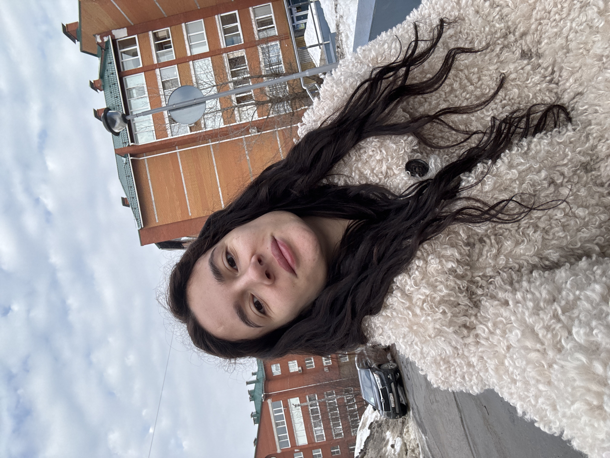 A female subject, approximately 20-30 years old, with long dark wavy hair and light skin, is depicted in a close-up selfie-style photograph. She is wearing a cream-colored, curly-pile textured coat. Her expression is neutral, with a slight upward curve to her lips.
The scene is an urban environment in Chaikovsky, Russia, during what appears to be late winter or early spring due to the presence of snow. In the background, to the left of the subject, a dark-colored Toyota Highlander SUV (Russian license plate X222AP 59) is parked on a paved street. Dirty snow banks are visible along the roadside. Behind the vehicle and extending to the right, multi-story residential buildings constructed of orange-red brick with green-tiled roofs and white-framed windows are visible. A grey streetlamp pole with a dark, enclosed light fixture and a circular blue-and-white road sign are positioned to the right of the center in the background. The sky is overcast with white and grey clouds.