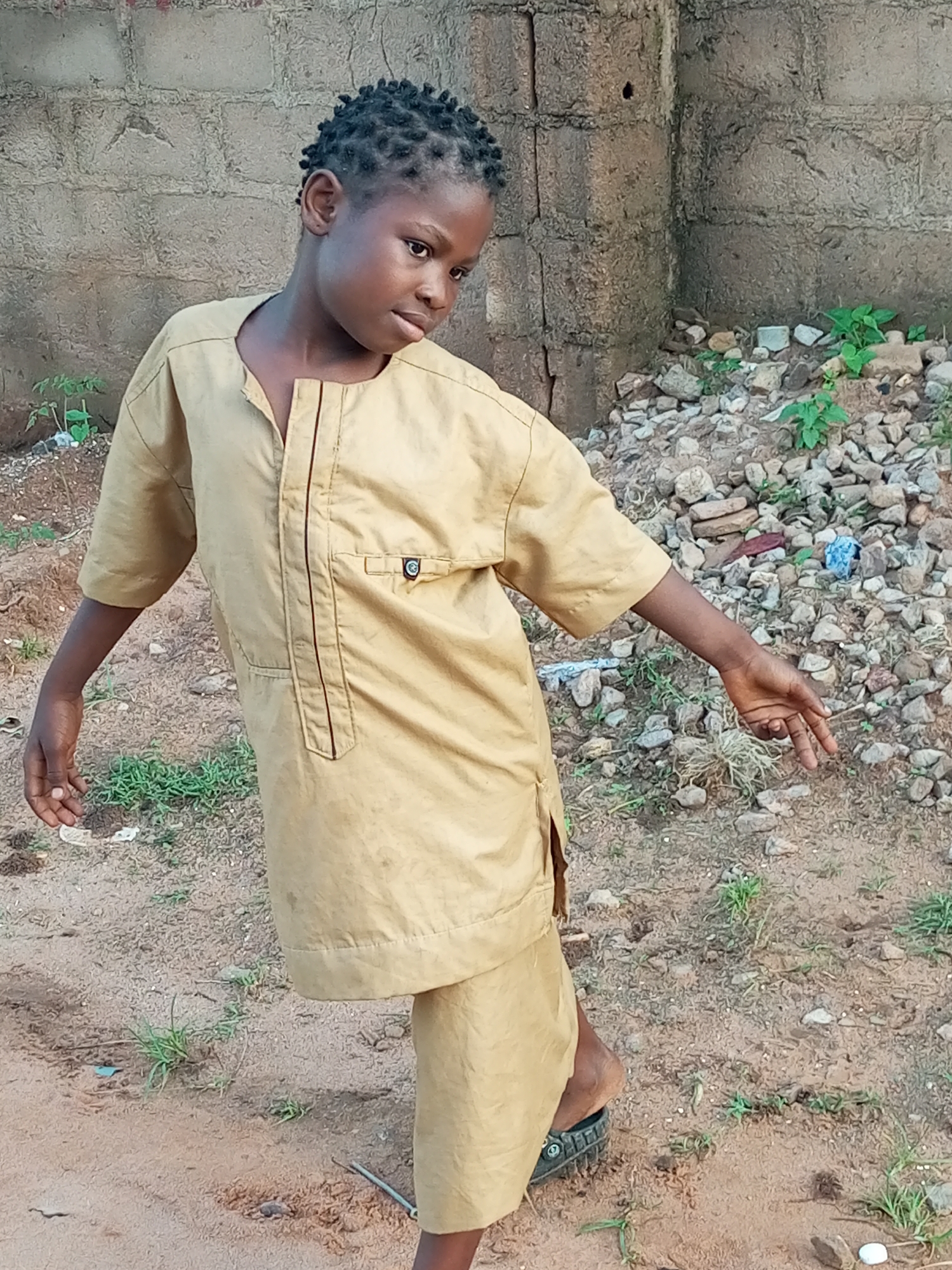 A young boy with dark skin and braided hair stands in a dirt area, facing towards the right of the frame. He is wearing a tan, loose-fitting garment with a V-neck opening and a pocket on the left side. The garment has a vertical seam down the front with a darker trim. His right arm is extended to his side, and his hand is open with fingers splayed. His left arm is also extended to his side, bent at the elbow, with his hand open. He is wearing dark sandals on his feet.
The background consists of a rough, grey brick wall on the left, and a pile of rocks and debris on the right. Small green plants are growing among the rocks and in the dirt. The ground is uneven and appears to be a mixture of dirt and gravel. The lighting suggests it is daytime, with the sun casting shadows. The location appears to be outdoors, possibly in a less-developed area, consistent with the city of Tempure, Nigeria.