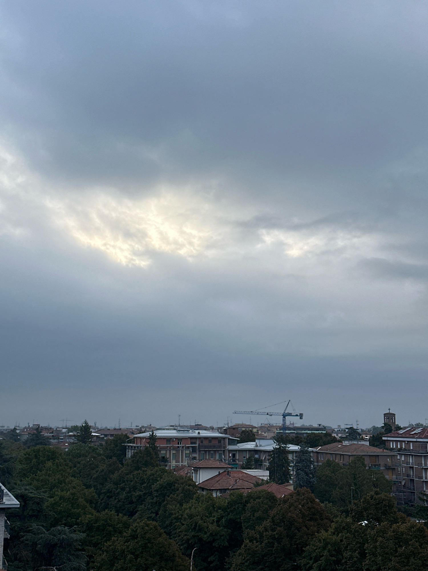 The image captures a cityscape under a cloudy sky, likely taken from an elevated position. The foreground is dominated by dense, dark green foliage, suggesting a park or wooded area within the city. Beyond the trees, a collection of buildings with varying architectural styles is visible. Some appear to be residential apartments, while others are larger, more institutional structures. A prominent blue construction crane stands out against the horizon, indicating ongoing development or infrastructure work in the area. In the distance, a church steeple can be seen, a common feature in Italian cities. The overall atmosphere is subdued, with the overcast sky casting a soft, diffused light on the scene. The location is Parma, Italy.