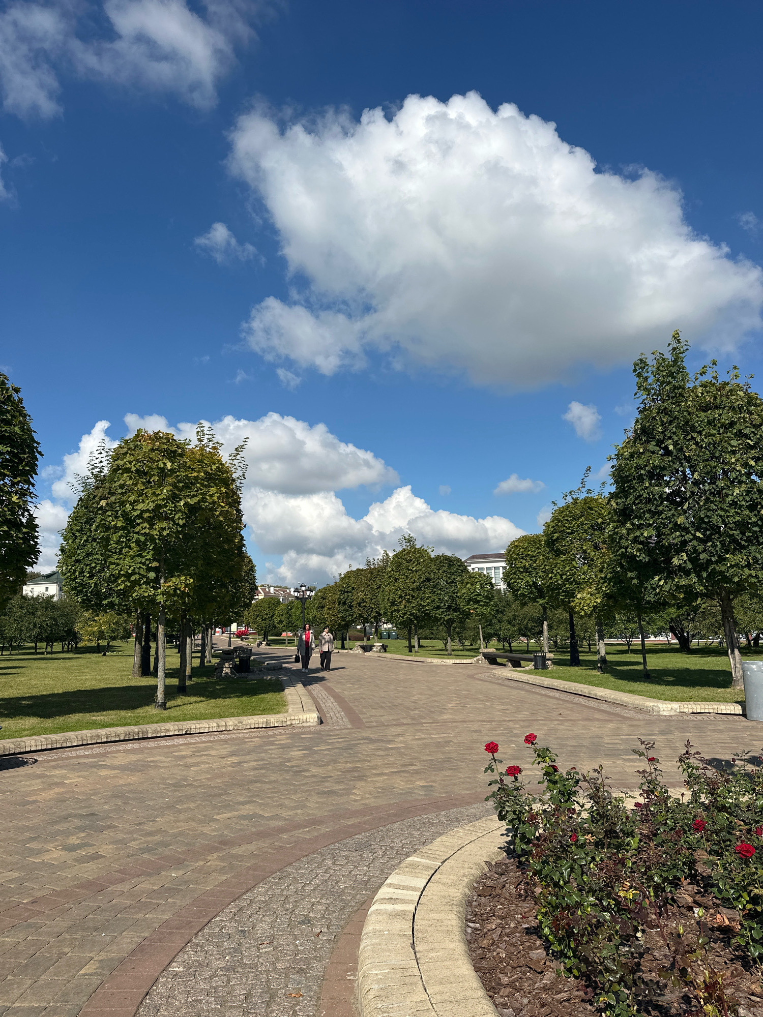 This is a photograph of a park in Minsk, Belarus, on a clear day with a few white clouds in a blue sky. The park features a paved walkway made of reddish-brown and grey interlocking bricks, with a curved border of light-colored stone. On the left side of the walkway, there is a grassy area with several neatly pruned trees. On the right side of the walkway, there are flower beds with red roses, followed by more trees and benches. In the distance, two people are walking away from the camera on the paved path. Beyond the trees, buildings with light-colored facades and roofs are visible. The overall impression is of a well-maintained, urban park.