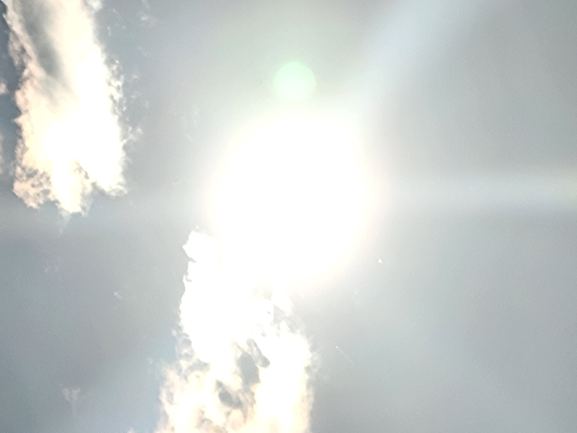 The image shows a bright sun shining through clouds, creating lens flare and a glowing effect. The sky is overcast with patches of bright white clouds. The sun is the dominant light source, casting a strong glare over the entire frame. There are no discernible people, subjects, or objects in the scene. The environment is an outdoor sky. The location is unknown.