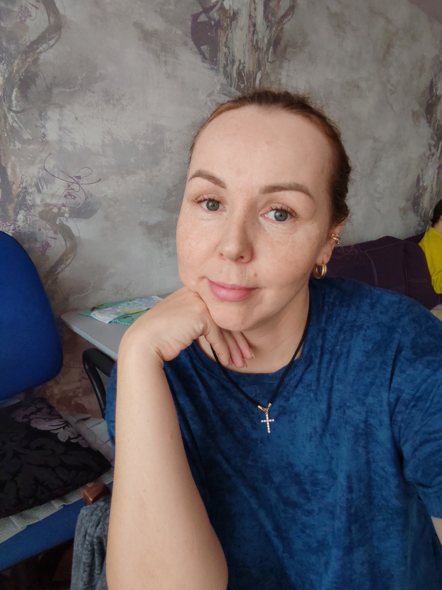 A medium shot shows a woman with light skin and red hair, wearing a blue t-shirt and a silver cross necklace, with her hand on her chin. She is looking at the camera with a neutral expression. In the background, a blue chair and a desk with papers are visible. A person in a yellow shirt is partially visible in the far right background. The wall behind the woman has a textured pattern. The scene appears to be indoors, likely a home office or study. The image does not contain any information about the city of Kizel, Russia.