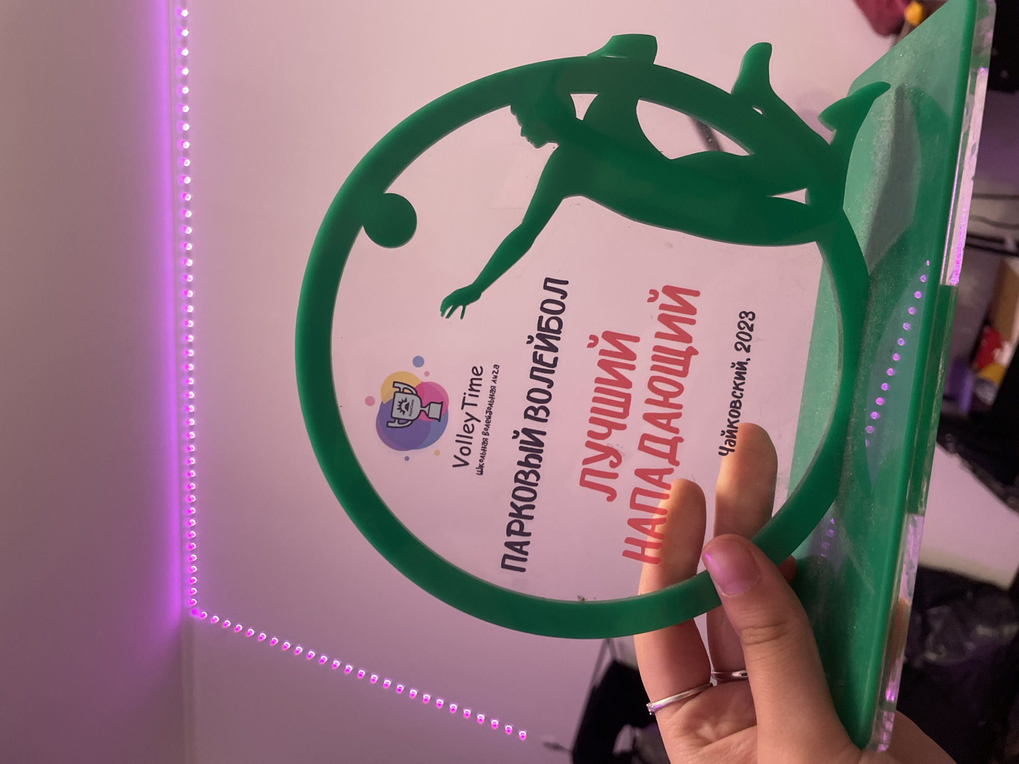 The image shows a trophy held by a person's hand in the city of Kokkorevo, Russia. The trophy is green and shaped like a volleyball player in motion, with a circular element behind. The text on the trophy reads "PARKOVY VOLEYBOL," "LUCHSHIY NAPADAYUSHCHIY," and "CHAYKOVSKIY, 2023." The background is a pink wall with purple LED strip lights along the ceiling. The person's hand is wearing a silver ring.
