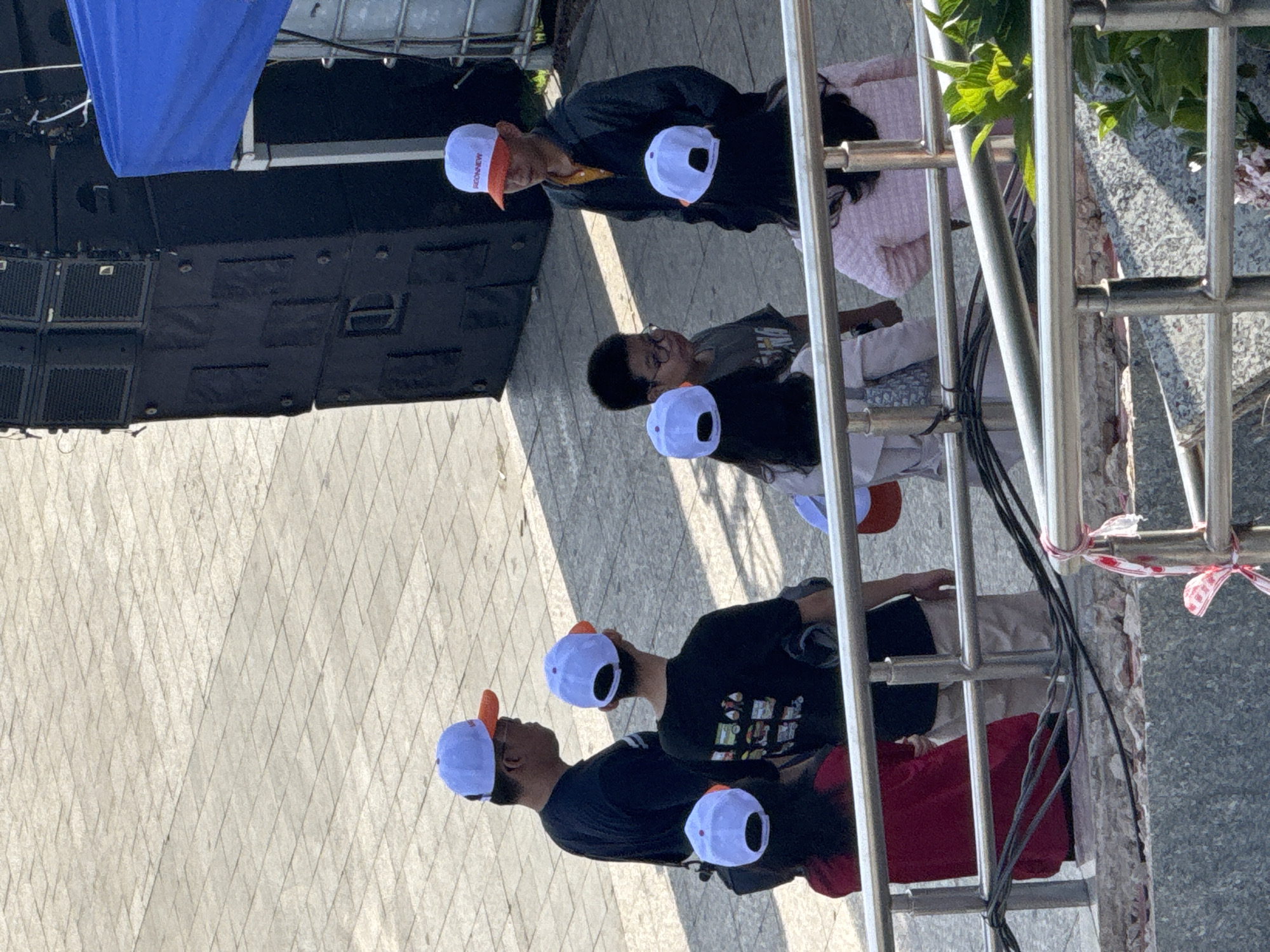In Da Lat, Vietnam, a group of people, predominantly wearing white and orange baseball caps, are gathered outdoors on a paved surface near a large black speaker system and a blue tarp. The scene appears to be a casual outdoor event or gathering. Several individuals are facing each other, engaged in conversation. One person in the foreground, wearing a red top and a white and orange cap, is partially visible. Behind them, a person in a black t-shirt with a graphic print and a white and orange cap is also present. A child with glasses stands to their right, looking forward. Further to the right, a young person in a dark jacket and a white and orange cap is speaking. Another individual in a pink textured jacket is also visible. The environment suggests a sunny day, with shadows cast on the ground. A metal railing runs across the lower portion of the image, obscuring some of the lower bodies of the people and the ground beneath them.
