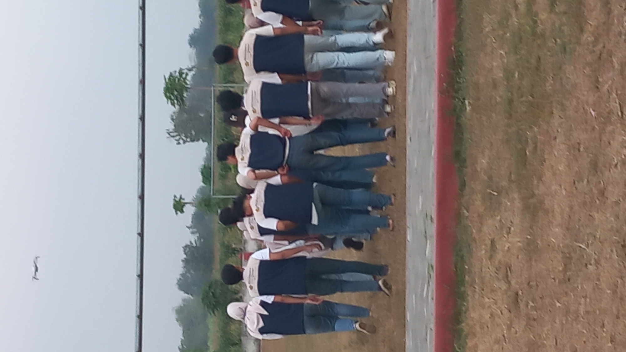A group of approximately 15 individuals, appearing to be young adults, are walking away from the camera in a field. They are dressed in matching uniforms consisting of dark blue vests over white shirts with light brown trim, and blue jeans. Several individuals are wearing headscarves. In the upper left corner of the frame, a small white drone is visible flying in the overcast sky. The environment appears to be an outdoor field, possibly a sports ground, with dry grass covering the ground. A metal fence or railing runs horizontally across the upper portion of the image. In the background, there are trees and a faint outline of a soccer goal. The setting is likely Pare, Indonesia, given the context.