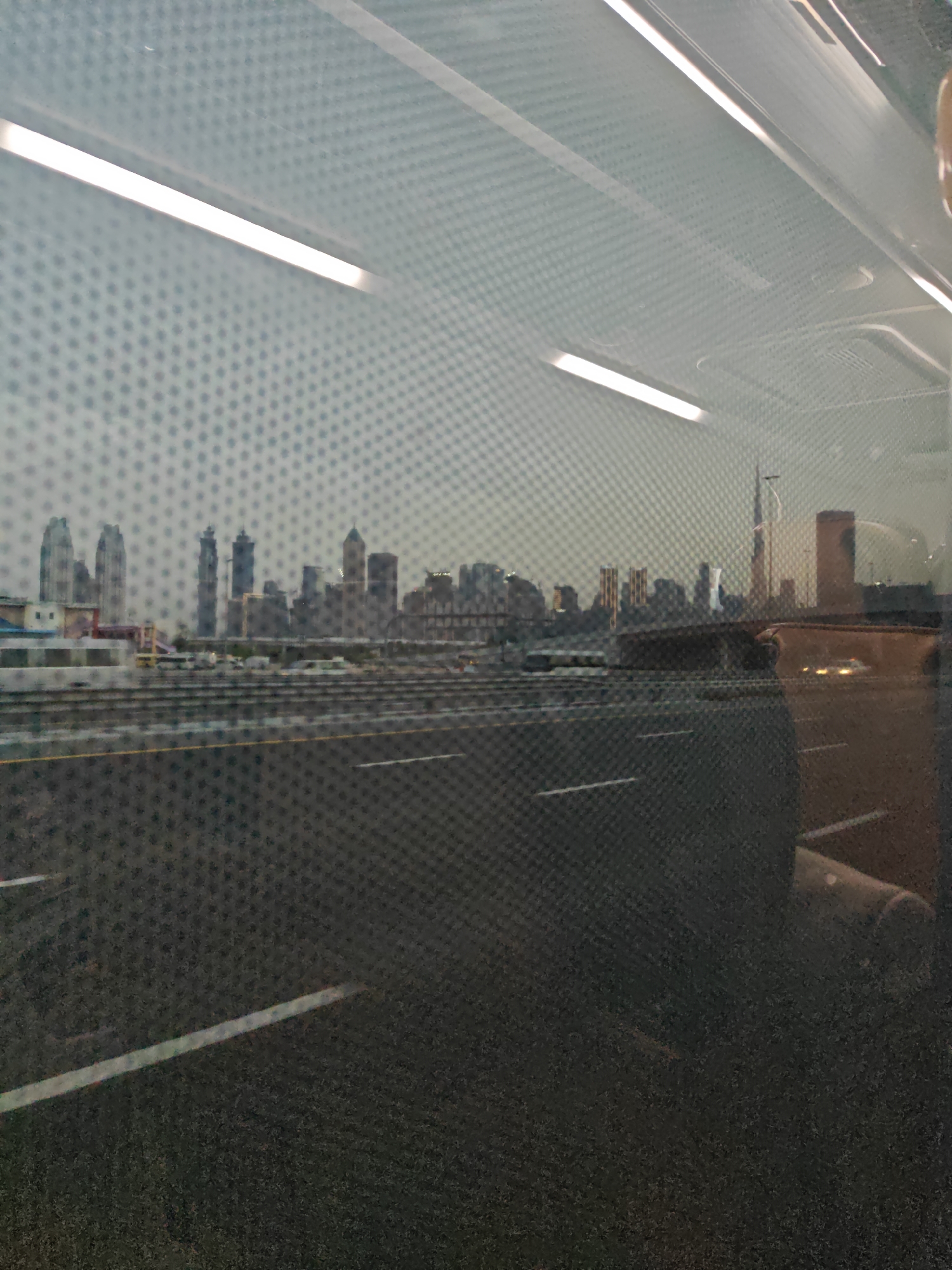 The image depicts a blurred view of a cityscape, likely Dubai, United Arab Emirates, seen through a textured surface, possibly a window with a patterned film or a rain-streaked surface. The foreground is dominated by a dark, asphalt-like surface with faint white lines, suggesting a road or a highway. In the mid-ground, a skyline of tall buildings is visible, characteristic of a major metropolitan area. Among these buildings, one distinctly resembles the Burj Khalifa, a prominent landmark in Dubai. There is also evidence of transportation infrastructure, with what appear to be train tracks or a metro line running horizontally across the scene. The sky is overcast, contributing to a muted color palette. Reflections on the surface obscure some details, adding to the overall impression of a distant or partially obscured view. The image appears to have been taken from a moving vehicle, as suggested by the slight motion blur.