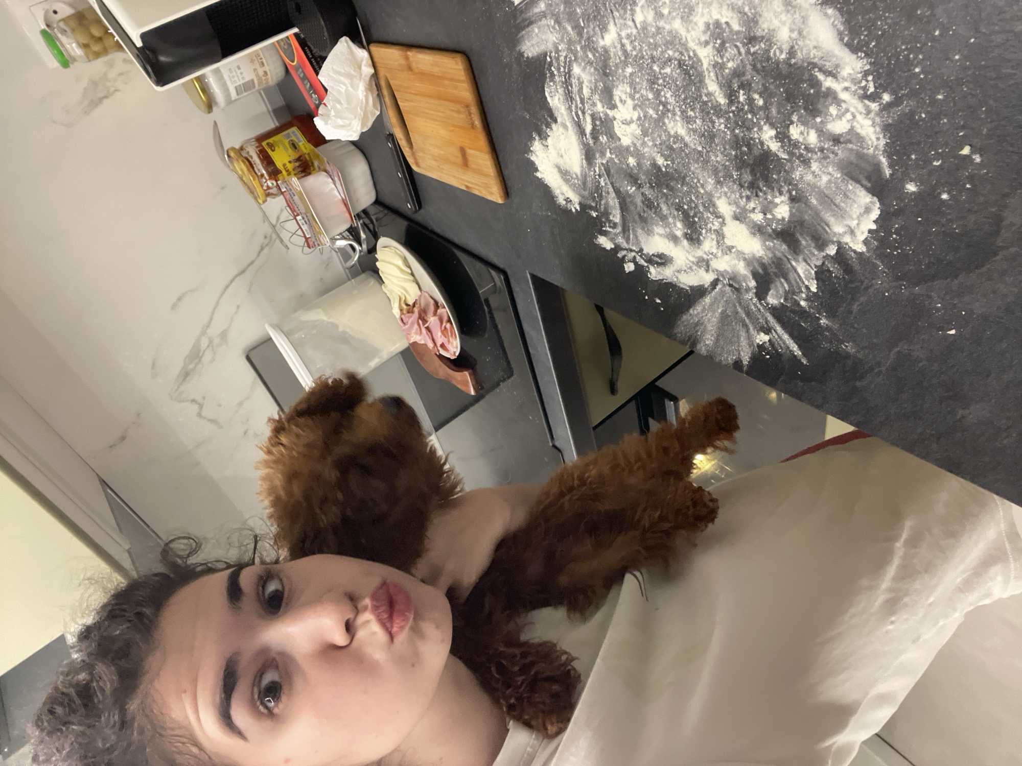 A woman with curly hair is holding a brown poodle close to her face. The scene appears to be inside a kitchen, with flour scattered on a dark countertop. Other items on the counter include a wooden cutting board, a knife, and a plate with what appears to be ham and cheese. Due to the lack of visual information, there is no means to determine the specific location within the city of Sofia, Bulgaria.