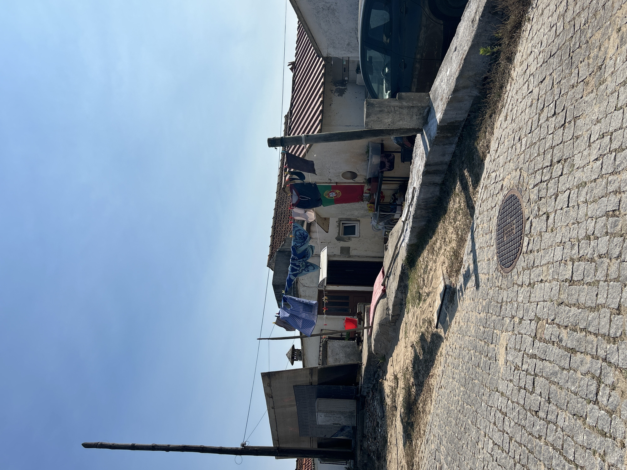The image depicts a rustic outdoor scene under a clear blue sky. In the foreground, a cobblestone pathway is visible, leading towards a low concrete wall. Beyond the wall, a sandy area with sparse vegetation is present. A cluster of simple, aged buildings with red-tiled roofs occupies the mid-ground. Clothes are hung on a line strung between a wooden pole and one of the buildings, suggesting everyday life. A Portuguese flag is displayed prominently on the facade of one of the structures. Various objects, including a red bucket and a rolled-up mat, are scattered around the ground. A dark-colored car is parked to the right of the buildings. A tall wooden pole stands to the left of the scene, with wires extending from it. The environment appears to be a rural or semi-rural setting, with a sense of informality.