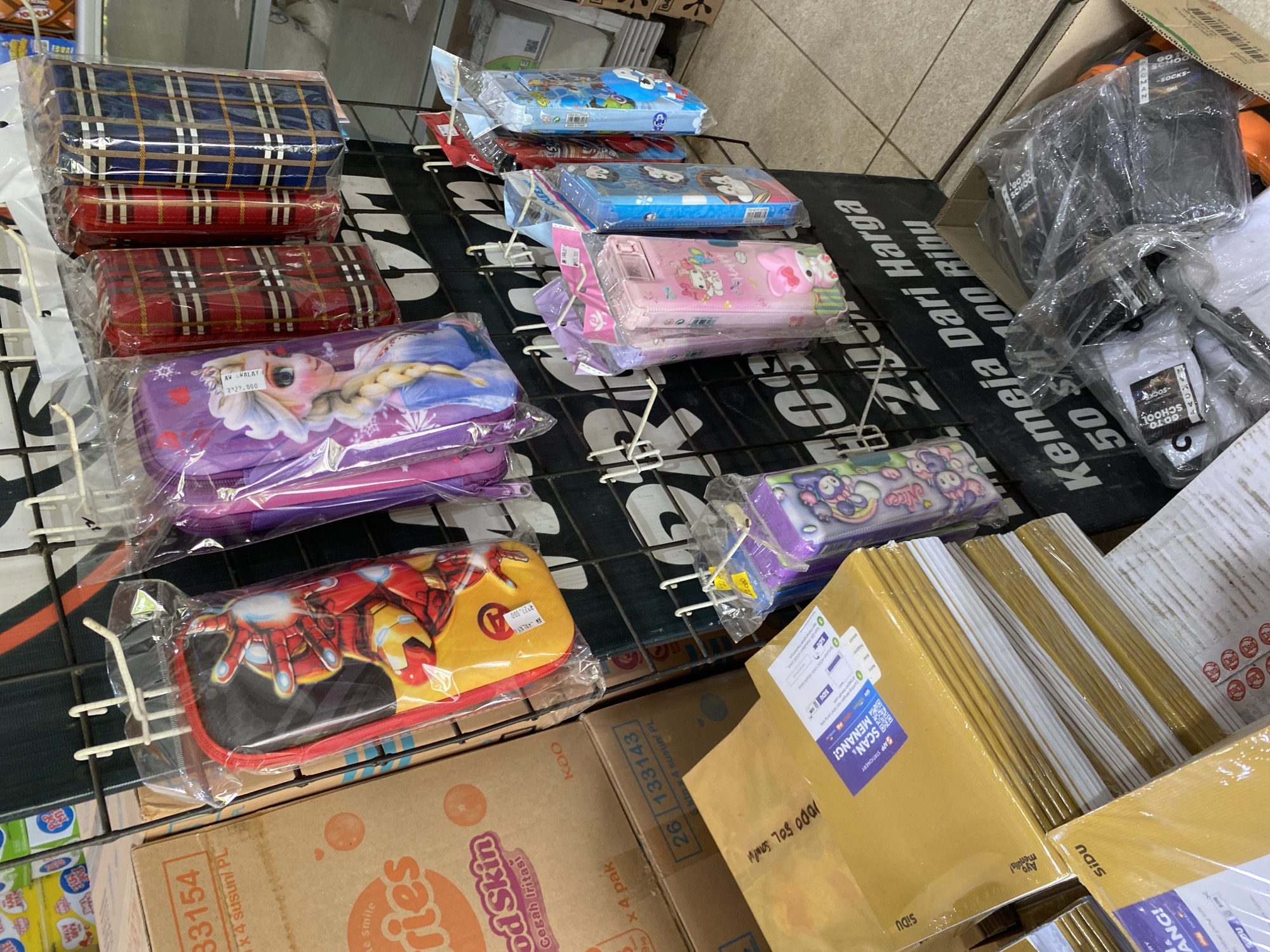 The image shows a retail display of stationery and other items in what appears to be a small shop or market stall. The items are organized on a black metal rack with white hooks.
In the foreground, there are stacks of yellow envelopes or folders, some with blue and white labels. To the left of these, part of a cardboard box is visible with text that includes "33154" and "Kao Good Skin".
The main focus of the display is on the rack. Hanging from the hooks are various pencil cases and pouches. These include a pencil case with an image of Iron Man, a purple pouch featuring Elsa from Frozen, and several other colorful pencil cases with cartoon characters. Further down the rack, there are more pencil cases, including pink ones with a Hello Kitty-like character and blue ones with a character resembling Doraemon.
To the right and slightly behind the main display, there is a cardboard box containing several pairs of packaged socks. Some of these are white with black text that says "100% CHOSOS", while others are black.
The background shows more shelving and merchandise, including what looks like snacks or packaged goods on shelves above the main display. The flooring appears to be tiled.
Based on the text visible on some of the products, such as "Sidu" on the yellow envelopes and "Kao" on the cardboard box, it is highly probable that this scene is located in Indonesia. The presence of these specific brands is common in Indonesian retail environments. Without more specific information or clearer signage in the image, it is impossible to pinpoint an exact location within Tanjong, Indonesia. However, the general context suggests a retail setting in that region.