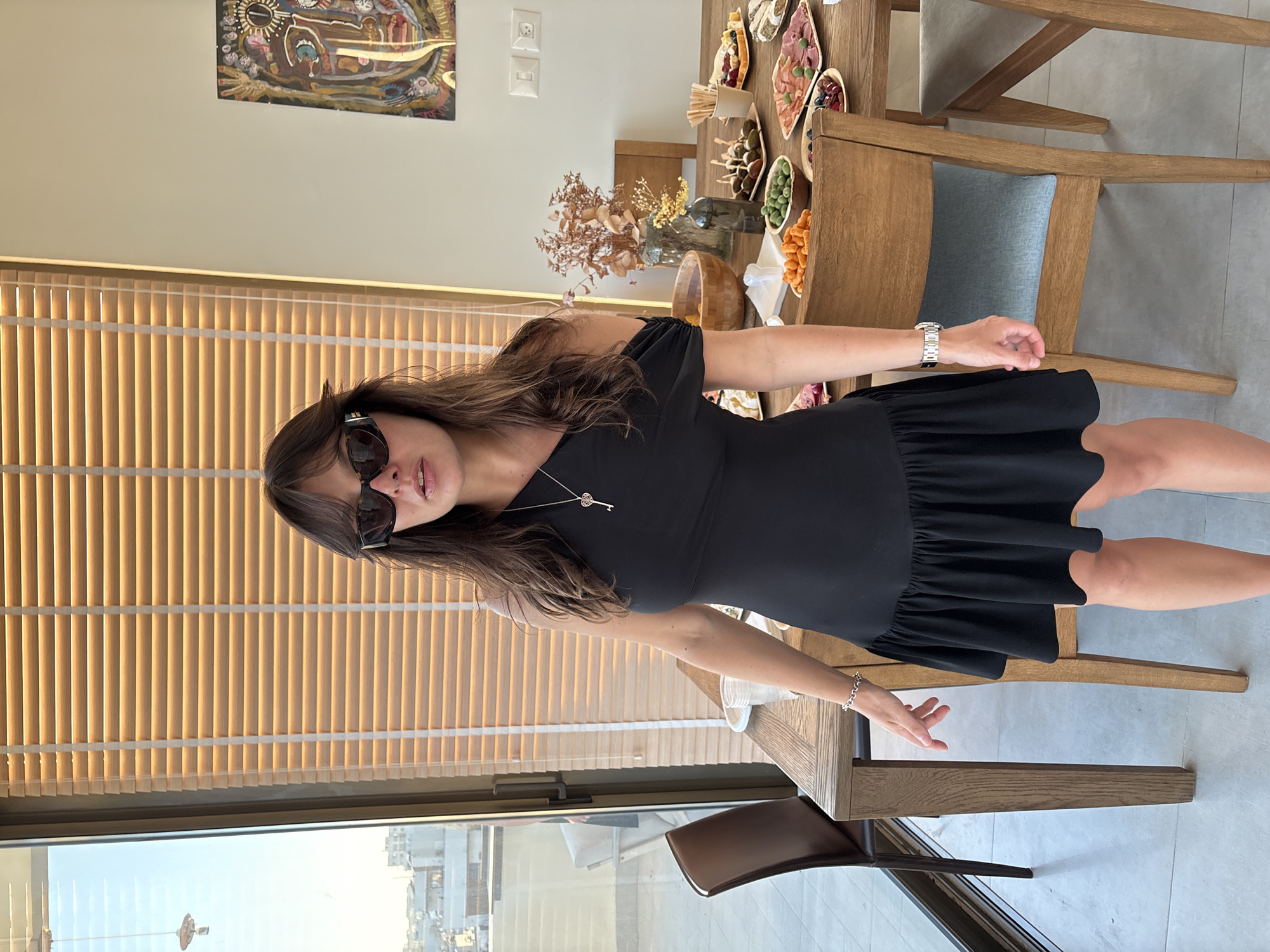A medium shot captures a woman standing in a dining area, likely in Tel-Aviv, Israel, as suggested by the distant cityscape visible through a large window with Venetian blinds. The woman, with long, dark, wavy hair and wearing large, dark sunglasses and a black, one-shoulder dress with a ruffled hem, stands facing the camera. She has a silver necklace with a key pendant and a silver wristwatch on her left wrist. Her left hand rests on her hip, while her right arm is slightly extended.
The woman is positioned near a wooden dining table laden with food platters, including various appetizers like olives, cheese, cured meats, and berries. A bouquet of dried flowers in a vase adds a decorative touch to the table. Several wooden chairs surround the table. In the background, a large abstract artwork hangs on the wall. The overall environment suggests a gathering or a meal preparation.