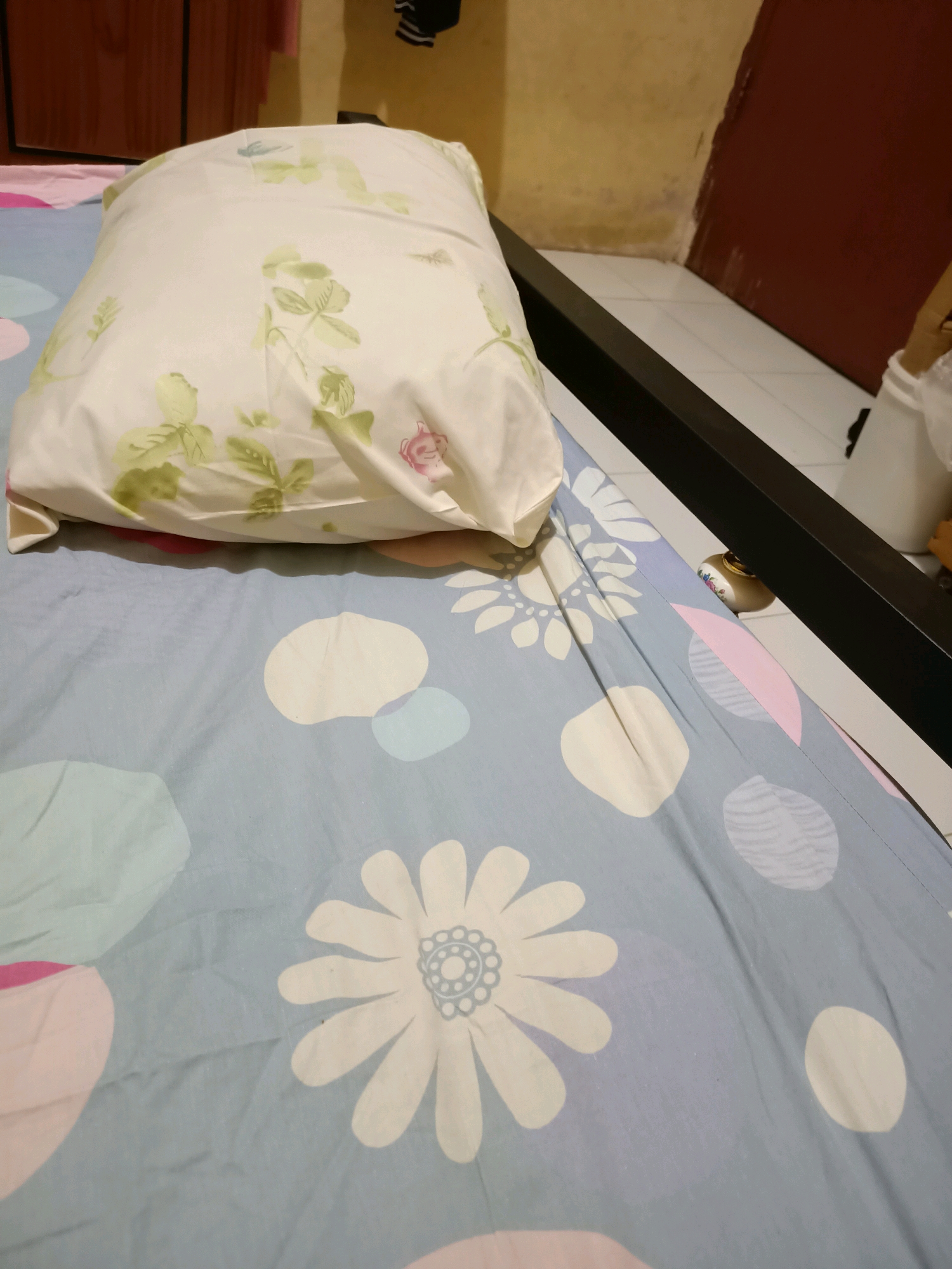 A close-up, eye-level shot of a bed with a pillow on it, taken indoors. The pillow has a white case with a light green floral pattern. The bedsheet is light blue with a pattern of white and pink abstract shapes and white flowers. The edge of a black metal bed frame is visible. In the background, a wall painted yellow and brown can be seen, with a white tiled floor. The overall scene suggests a domestic bedroom setting, likely in a residential area of Perawang, Indonesia. No people or explicit actions are depicted.