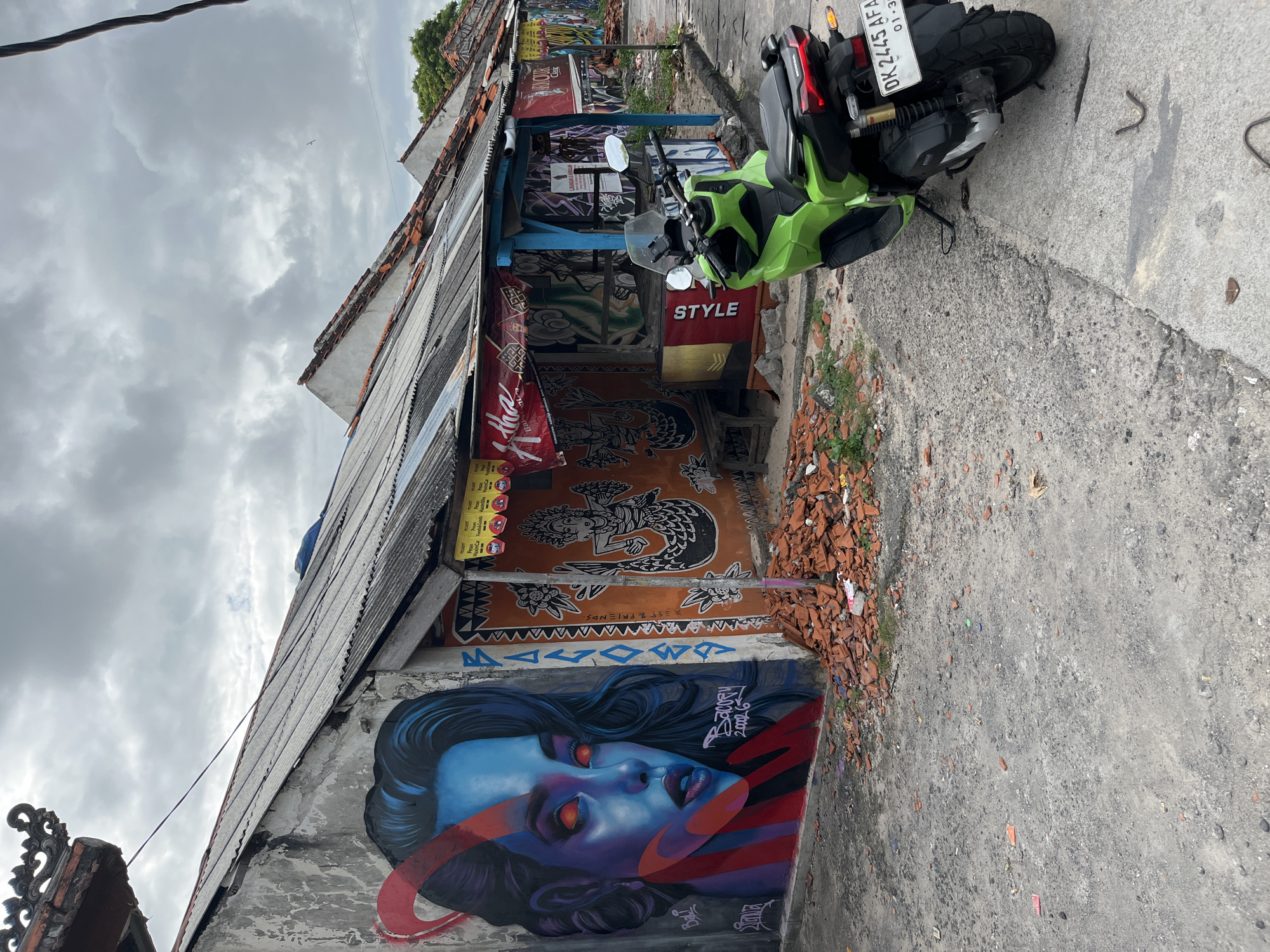 The image shows a street scene in Kuta Selatan, Indonesia, featuring a row of buildings adorned with colorful murals and graffiti. On the left, a large mural of a woman with blue skin and glowing red eyes dominates the wall of a building. To the right, the buildings have corrugated metal roofs and are covered in various artistic expressions, including a depiction of a mermaid and intricate patterns. A bright green scooter is parked on the asphalt road in front of one of the shops. Debris, appearing to be broken terracotta tiles, is scattered on the ground near the base of the buildings. The sky is overcast with gray clouds. The overall impression is one of an urban environment with a vibrant street art culture.