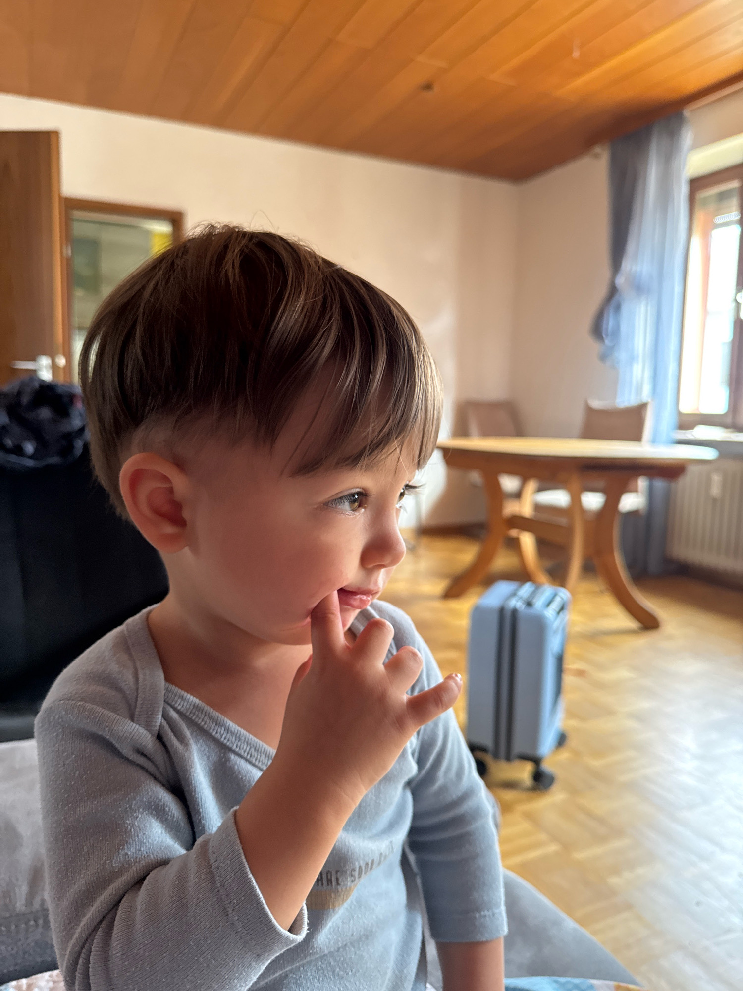 In a domestic indoor setting, a young child is the central focus, viewed in profile from the left. The child has brown hair and is wearing a light blue long-sleeved shirt. Their right index finger is placed near their mouth. The background shows a wooden ceiling, a wall, a window with blue curtains, and a wooden table with chairs. A small blue suitcase is visible on the floor in the middle ground. The overall scene suggests a casual moment within a home.