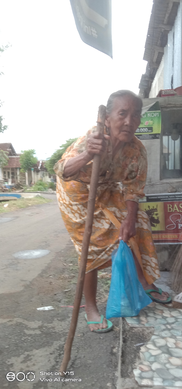 An elderly woman, with grey hair pulled back, stands on a concrete curb beside an unpaved road in Maron, Indonesia. She wears a yellow and white batik-patterned dress and light green flip-flops. She leans on a rough, unrefined wooden walking stick held in her right hand and carries a translucent blue plastic bag in her left hand. Her gaze is directed towards the viewer. The road to her left exhibits several small puddles, and in the background, multiple houses with reddish-brown tiled roofs and white walls are visible among sparse trees. To her right, a building's facade features a green banner advertising "KOPI" with a price, and a brown sign with a crown logo for "BASO" and "KRETEK" cigarettes. A red box marked "20271" is affixed to the building's upper right, and a dark banner with white, reflective text hangs partially overhead. The sky is bright and appears overcast.