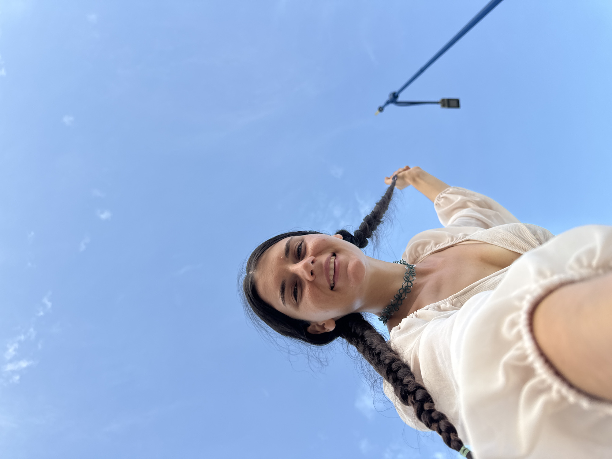 A low-angle upward shot captures a young woman against a clear blue sky with sparse white clouds. The woman, with long dark hair styled in two braids, looks directly at the camera with a wide, open-mouthed smile, conveying a sense of happiness. She is wearing a light-colored, possibly cream or white, off-the-shoulder top with ruffled sleeves and a dark green, intricately patterned choker necklace. Her right hand is visible, holding one of her braids playfully. In the upper right corner of the frame, a dark blue bar or cable extends diagonally across the sky, with a dark, rectangular street light visible beneath it, suggesting an outdoor urban or semi-urban environment. The bright, even lighting indicates it is daytime. The overall impression is a joyful moment captured on a clear day, with the context being Mui Ne, Vietnam. There is no visible text.