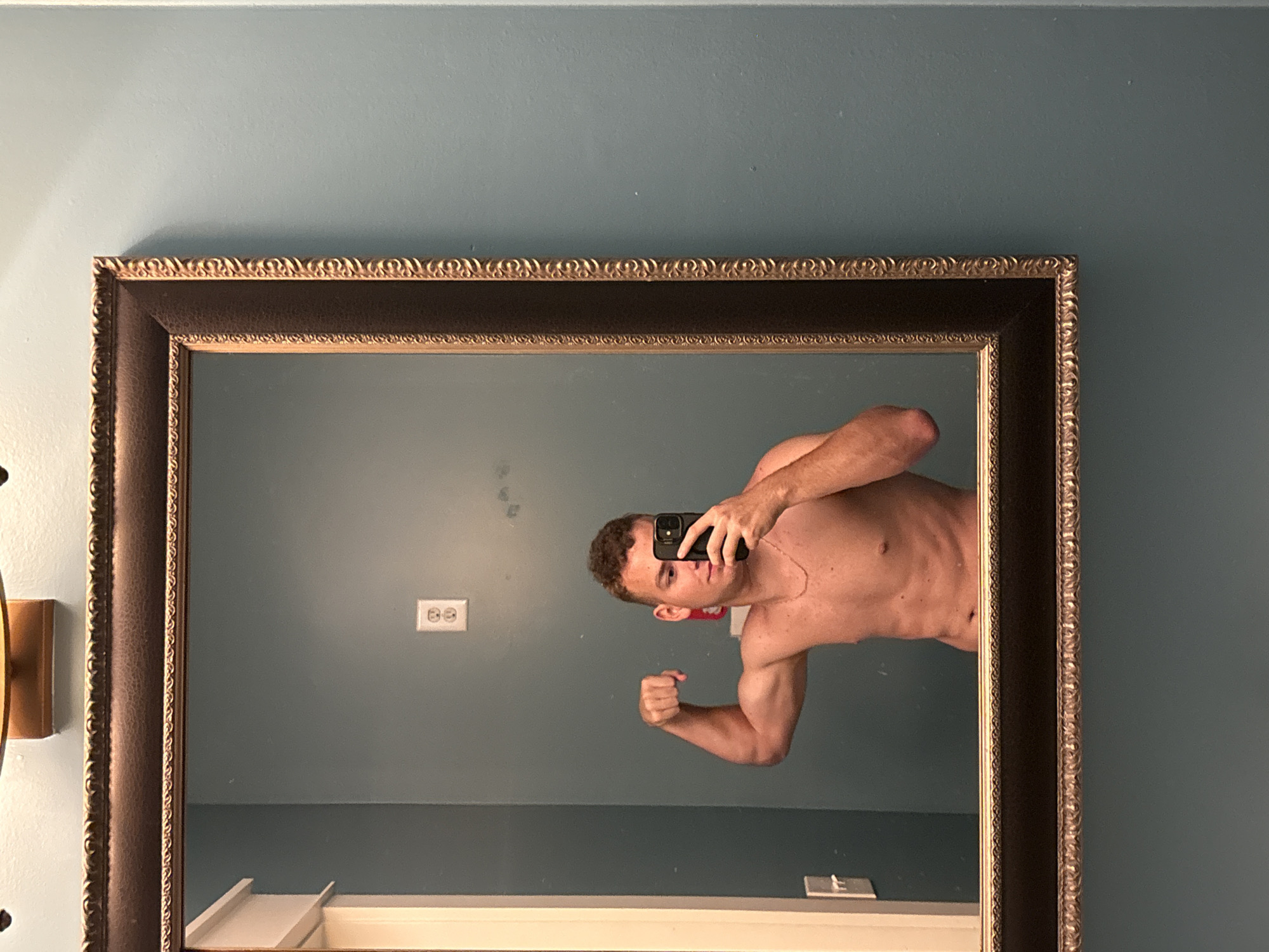 A shirtless man is taking a mirror selfie in a bathroom. He is standing in front of a teal wall, with a dark framed mirror. He is flexing his bicep on the left side of the image. He is holding a phone in his right hand, which is positioned to take the picture. The room appears to be a residential bathroom, possibly in a hotel or rental property, located in Panama City Beach, Florida, United States of America, as indicated by the context of the image being a reflection of a person in a bathroom, commonly found in such locations.