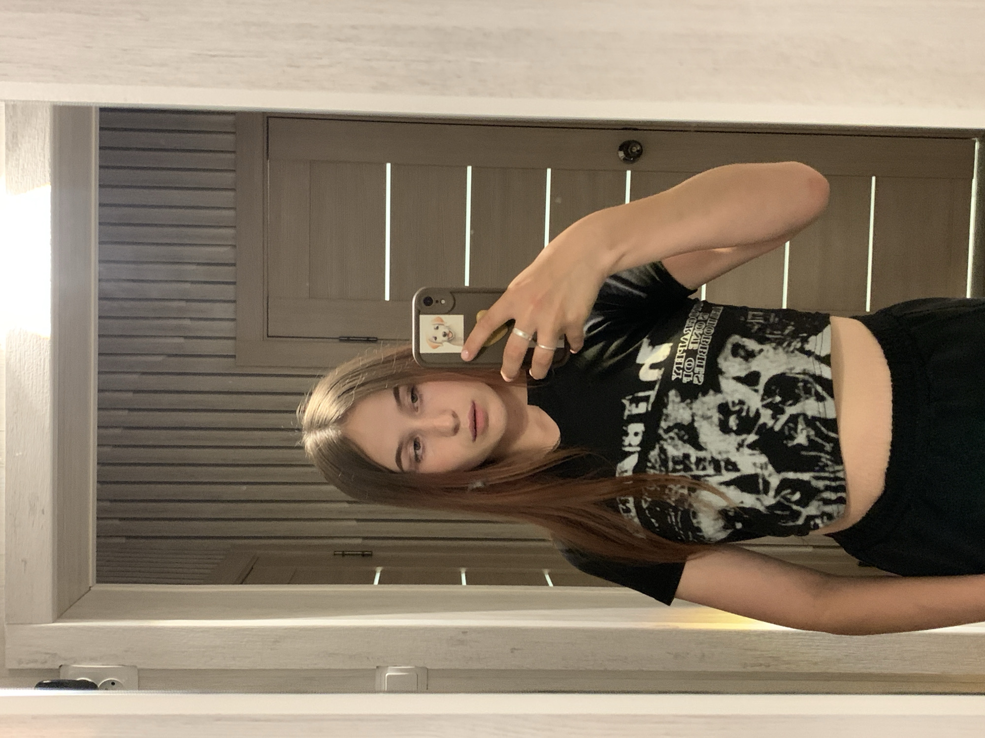 A young woman takes a selfie in a mirror indoors. She has long brown hair and is wearing a black crop top. The background includes a wall with horizontal stripes and a wooden door. A phone with a sticker on the back is visible in her hand. The location is not discernible from the image to be Фоки, Russia.